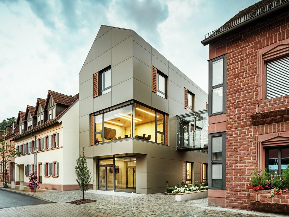 La photo montre la mairie moderne d'Alzenau, revêtue de panneaux composites en aluminium PREFA dans un ton bronze chaleureux. La façade est interrompue par de grandes surfaces vitrées qui offrent une vue sur l'intérieur, éclairé par une lumière chaude. La passerelle vitrée à droite de l'image relie le bâtiment à un bâtiment traditionnel en briques situé à proximité, créant ainsi un contraste intéressant entre l'architecture moderne et historique.