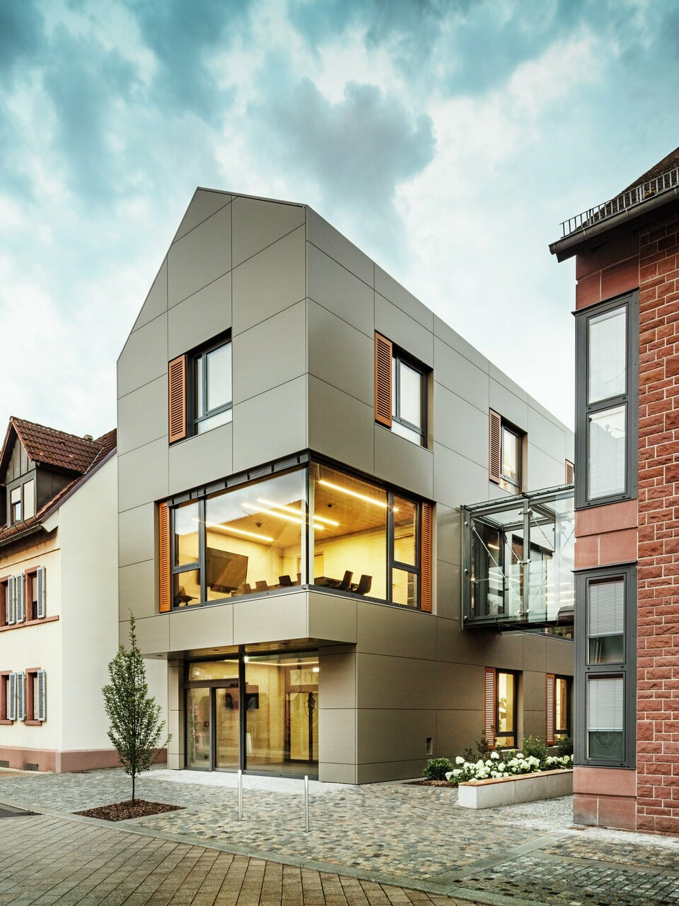 La photo montre la mairie moderne d'Alzenau, revêtue de panneaux composites en aluminium PREFA dans un ton bronze chaleureux. La façade est interrompue par de grandes surfaces vitrées qui offrent une vue sur l'intérieur, éclairé par une lumière chaude. La passerelle vitrée à droite de l'image relie le bâtiment à un bâtiment traditionnel en briques situé à proximité, créant ainsi un contraste intéressant entre l'architecture moderne et historique.