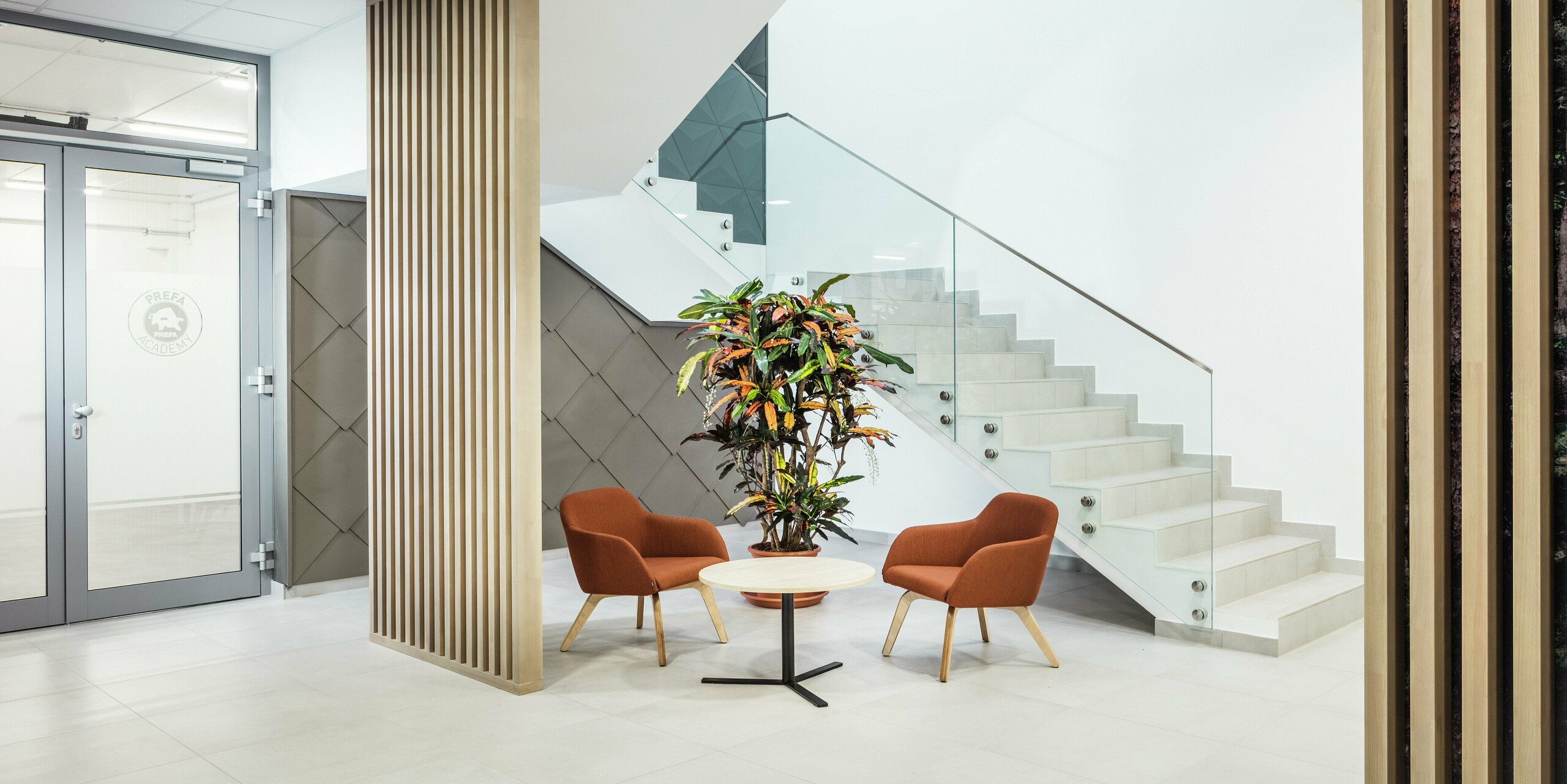 [Translate to Français:] Das stilvolle Interieur des neuen PREFA Standortes in Törökbálint (Ungarn) überzeugt durch eine moderne und einladende Gestaltung. Neben dem Eingang zu den praktischen Schulungsräumen der PREFA Academy wurde die Wandverkleidung mit PREFA Wandrauten 44 × 44 in Bronze akzentuiert. Der Eyecatcher zieht sich von der Außenfassade bis ins Innere und verbindet dabei Design mit Funktionalität. Auch im Innenbereich fügt sich die hochwertige Aluminiumoberfläche harmonisch in das Gesamtkonzept ein. Neben hellen Holzelementen wurden einladende Sitzmöbel und Grünpflanzen platziert, um den Zugang zu den Büros über das Treppenhaus für die Mitarbeiterinnen und Mitarbeiter angenehmer zu gestalten.