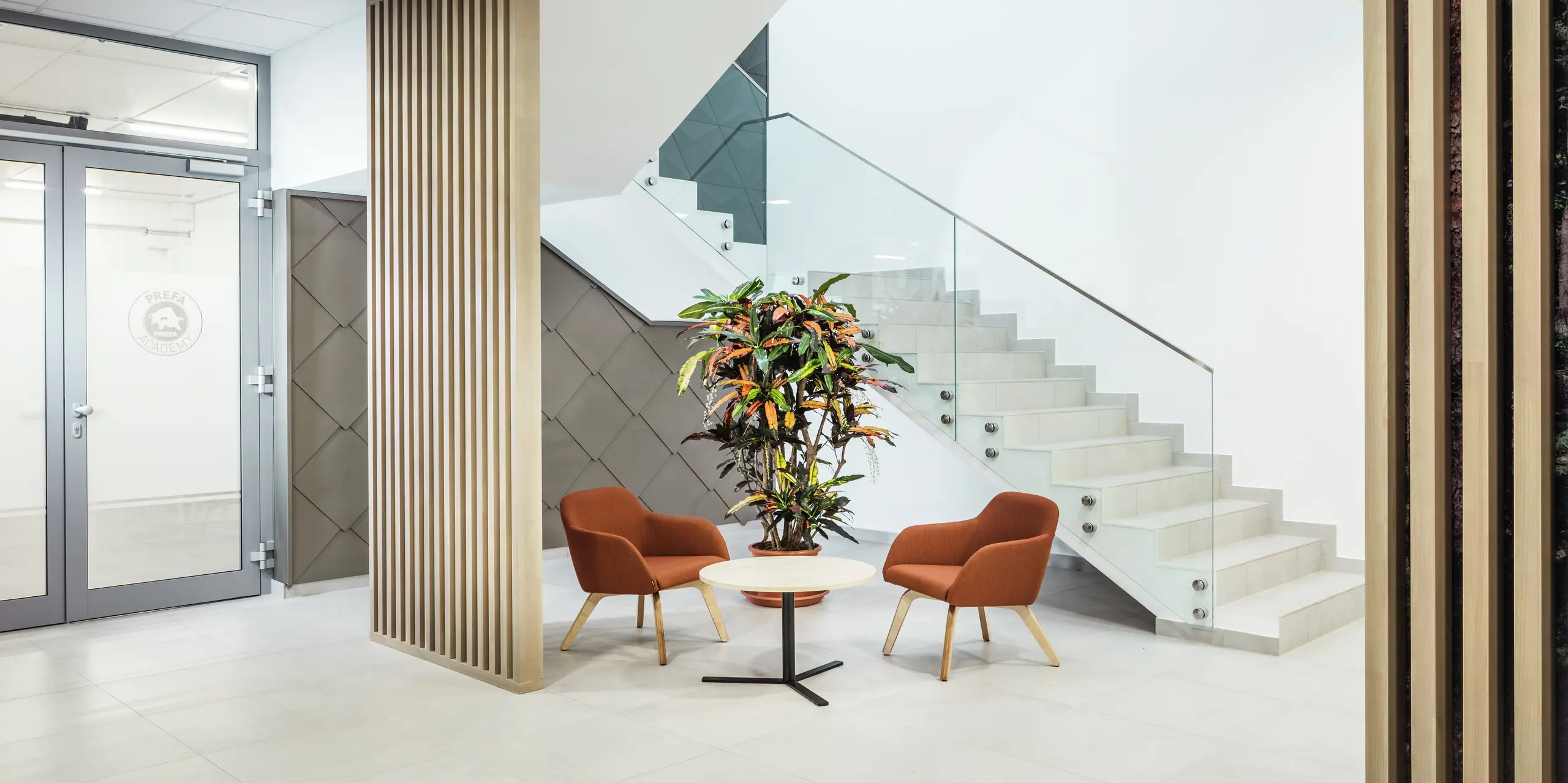 [Translate to Français:] Das stilvolle Interieur des neuen PREFA Standortes in Törökbálint (Ungarn) überzeugt durch eine moderne und einladende Gestaltung. Neben dem Eingang zu den praktischen Schulungsräumen der PREFA Academy wurde die Wandverkleidung mit PREFA Wandrauten 44 × 44 in Bronze akzentuiert. Der Eyecatcher zieht sich von der Außenfassade bis ins Innere und verbindet dabei Design mit Funktionalität. Auch im Innenbereich fügt sich die hochwertige Aluminiumoberfläche harmonisch in das Gesamtkonzept ein. Neben hellen Holzelementen wurden einladende Sitzmöbel und Grünpflanzen platziert, um den Zugang zu den Büros über das Treppenhaus für die Mitarbeiterinnen und Mitarbeiter angenehmer zu gestalten.