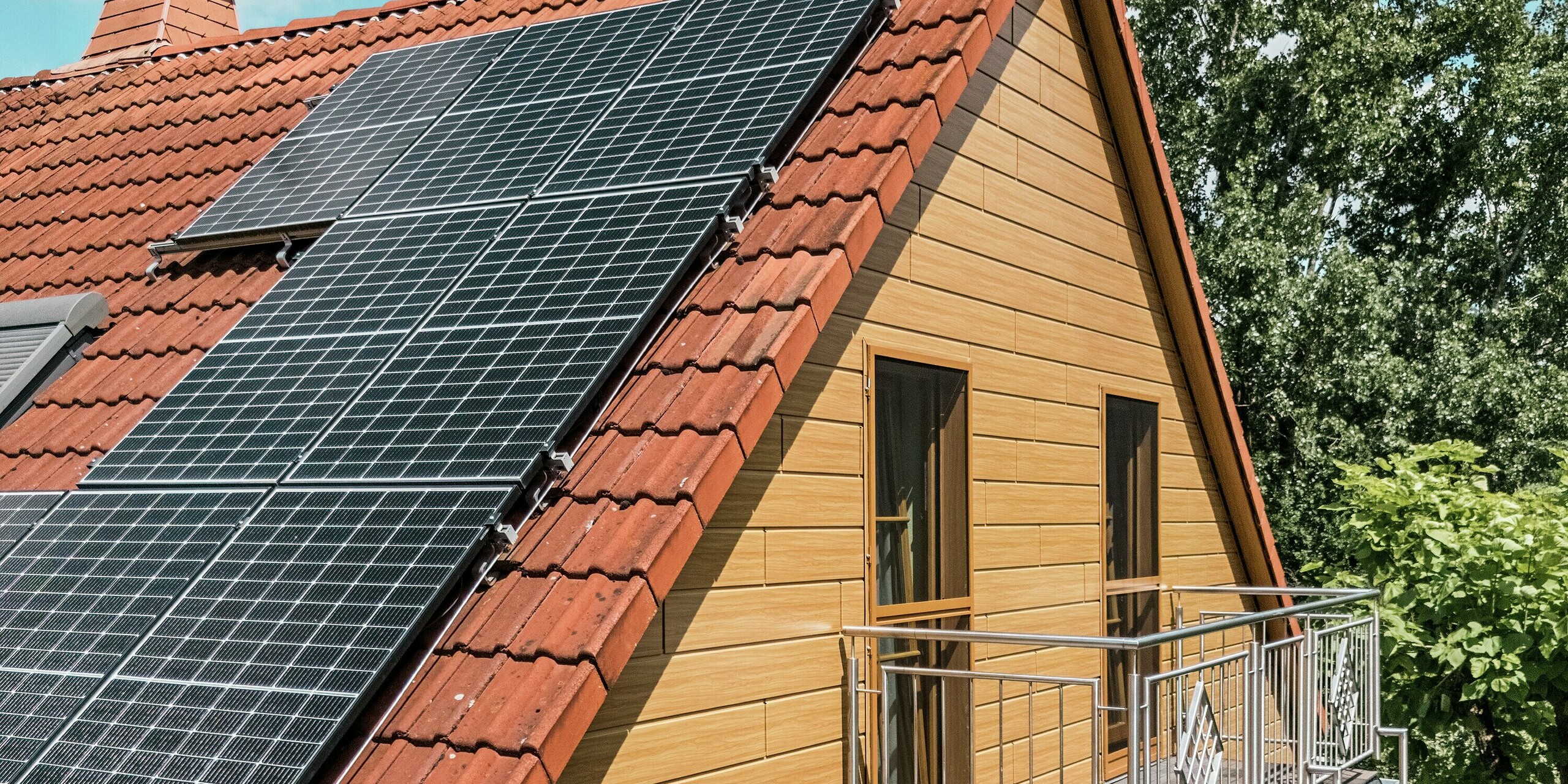 L'image présente le pignon d'une maison individuelle à Blieskastel, rénové avec goût et revêtu de Siding PREFA en chêne naturel. L'élégante façade en aluminium, à l'aspect bois réaliste, crée un contraste chaleureux avec le rouge éclatant de la toiture en tuiles et les grands modules photovoltaïques qui scintillent au soleil. Le balcon délicat, avec sa rambarde métallique, souligne les lignes élégantes de la maison. Grâce à la surface PREFA résistante aux intempéries et inaltérable, l'aspect bois de haute qualité est préservé durablement : un atout esthétique et une protection longue durée.