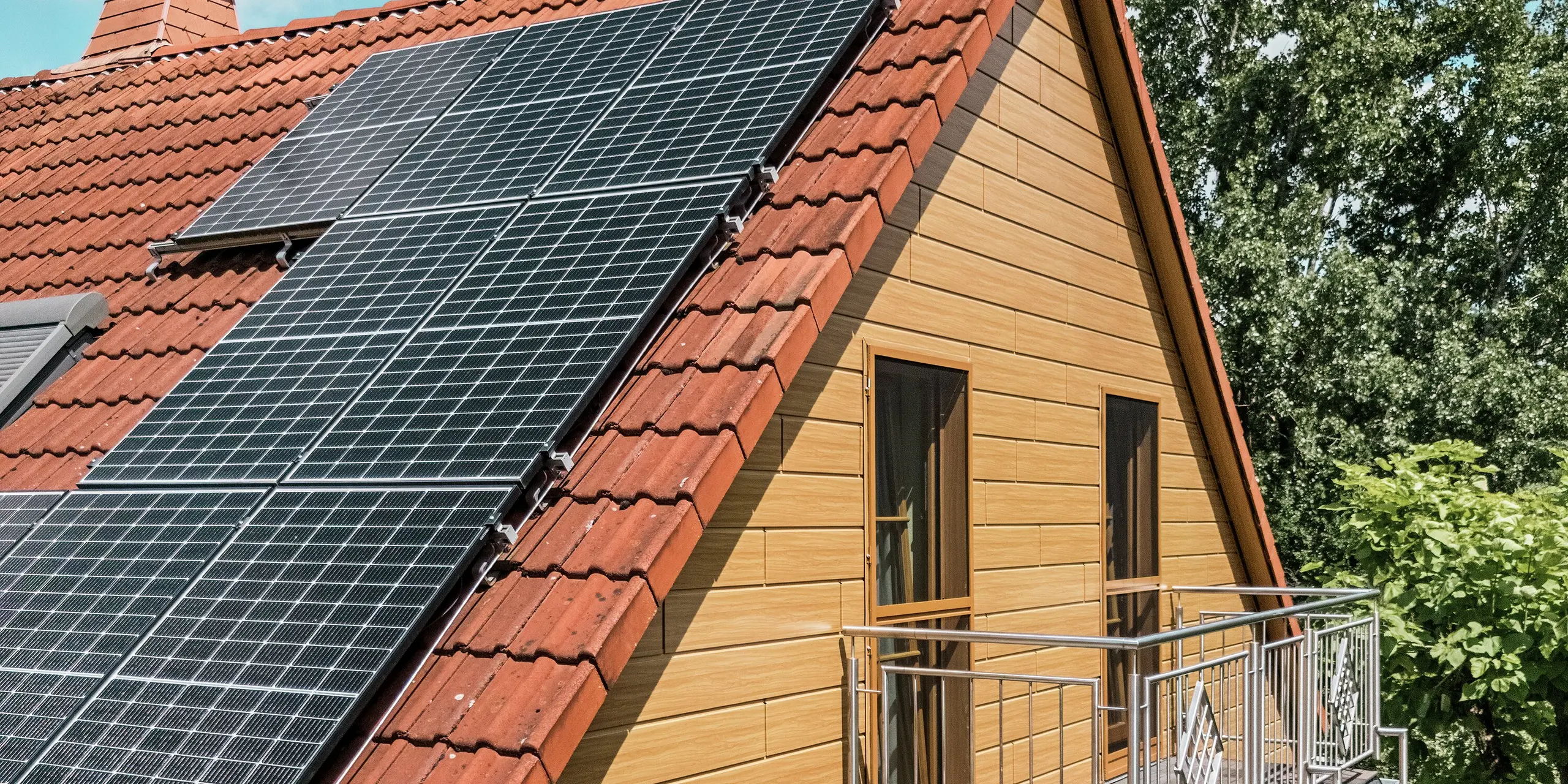 L'image présente le pignon d'une maison individuelle à Blieskastel, rénové avec goût et revêtu de Siding PREFA en chêne naturel. L'élégante façade en aluminium, à l'aspect bois réaliste, crée un contraste chaleureux avec le rouge éclatant de la toiture en tuiles et les grands modules photovoltaïques qui scintillent au soleil. Le balcon délicat, avec sa rambarde métallique, souligne les lignes élégantes de la maison. Grâce à la surface PREFA résistante aux intempéries et inaltérable, l'aspect bois de haute qualité est préservé durablement : un atout esthétique et une protection longue durée.