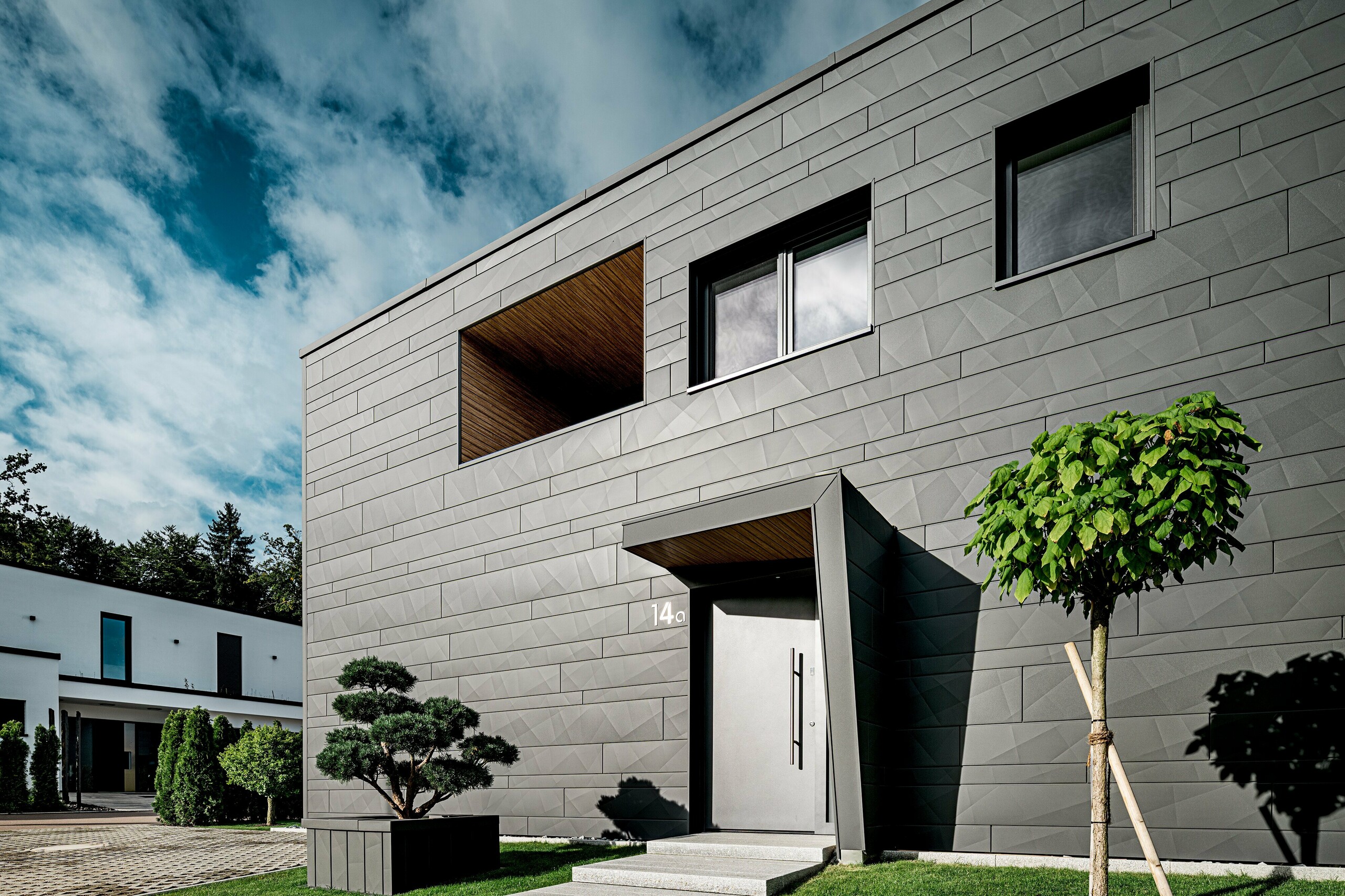 Das moderne Einfamilienhaus in Heidenheim präsentiert sich mit einer markanten PREFA Siding.X Fassade in P.10 Dunkelgrau, deren kantige Struktur das klare Design betont. Der Eingang mit tiefem, holzverkleidetem Rücksprung erzeugt einen spannenden architektonischen Fokuspunkt. Im Vordergrund setzen ein geschnittener Formbaum und ein junger Kugelbaum lebendige Akzente. Die widerstandsfähige Aluminiumfassade sowie das integrierte PREFA Quadratrohr sorgen für dauerhaften Schutz, moderne Optik und ein hochwertiges Gesamtbild.