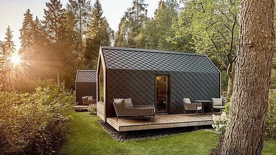 Nichée dans un écrin de verdure paisible au bord du lac Natterer, une minuscule maison du Secret Koi Garden est baignée par la douce lumière du soleil couchant. Son toit et sa façade sont revêtus de petits panneaux d'aluminium PREFA en forme de losange, couleur P.10 anthracite, dont la texture évoque la peau d'un poisson. Robuste et durable, la structure en aluminium offre une protection efficace contre les intempéries et souligne l'architecture épurée et intemporelle de ce glamping en pleine nature.
