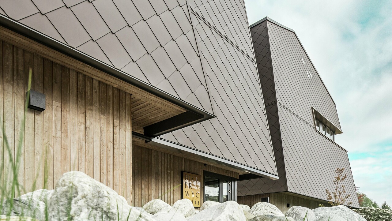 Vue latérale d'un hôtel, la façade du rez-de-chaussée est revêtue verticalement de bois plutôt sombre. Au-dessus se trouve un avant-toit, à partir duquel la façade est revêtue de plaques d'aluminium brun noisette.