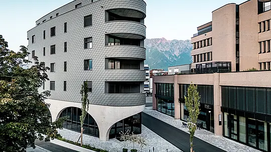 Vue de la Bernerhaus à Buchs, avec sa forme triangulaire remarquable et ses balcons aux courbes douces se détachant sur un fond de montagnes alpines. La façade continue en bardage PREFA gris souris P.10 enveloppe le bâtiment comme une peau d'aluminium aux allures d'écailles, créant un jeu d'ombre et de lumière saisissant. Au-dessus du socle en béton massif aux élégantes arcades, la façade légère et résistante aux intempéries en aluminium PREFA se distingue par sa précision, sa durabilité et son faible besoin d'entretien – un véritable atout architectural dans le paysage urbain.
