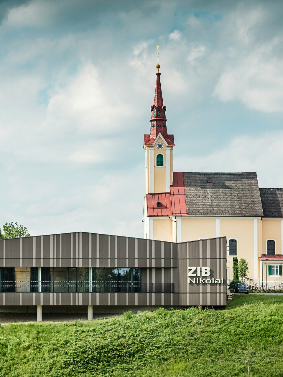 Le centre événementiel ZIB Nikolai à St. Veit dans le sud de la Styrie avec une façade en aluminium moderne et dynamique dans des tons gris brun-beige, au premier plan une prairie verte, à l'arrière-plan l'église Saint-Nicolas avec une façade jaune et un toit rouge.