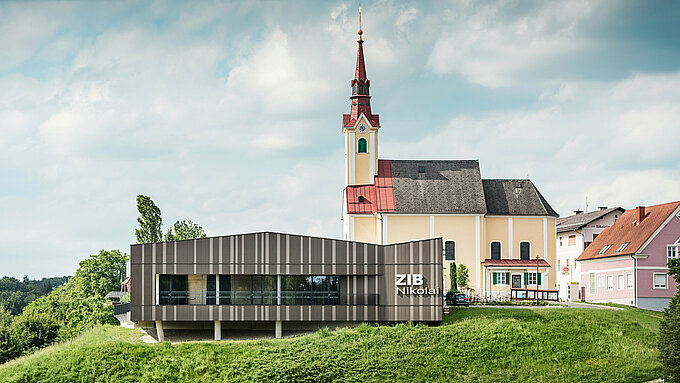 Le centre événementiel ZIB Nikolai à St. Veit dans le sud de la Styrie avec une façade en aluminium moderne et dynamique dans des tons gris brun-beige, au premier plan une prairie verte, à l'arrière-plan l'église Saint-Nicolas avec une façade jaune et un toit rouge.