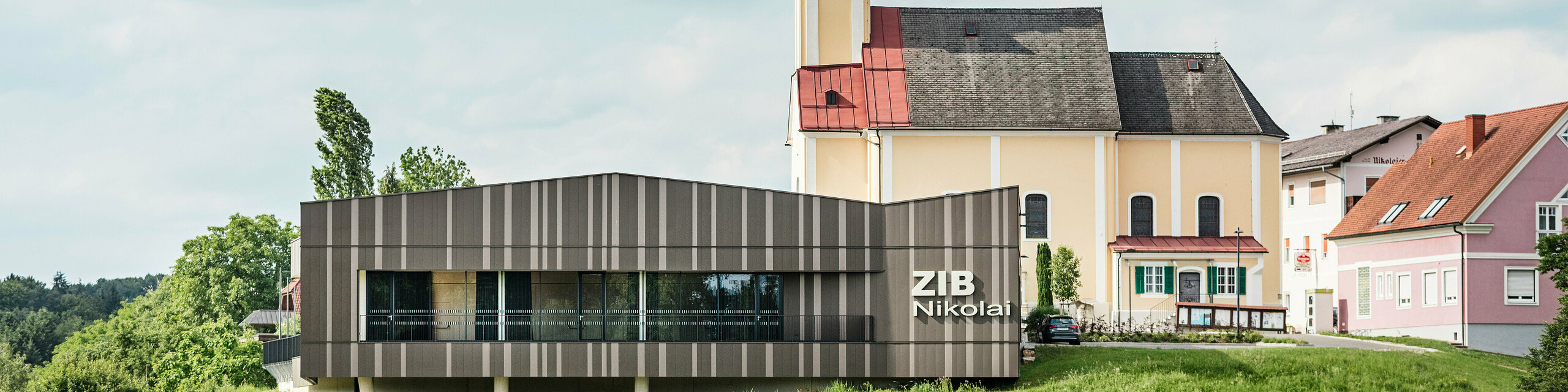 Le centre événementiel ZIB Nikolai à St. Veit dans le sud de la Styrie avec une façade en aluminium moderne et dynamique dans des tons gris brun-beige, au premier plan une prairie verte, à l'arrière-plan l'église Saint-Nicolas avec une façade jaune et un toit rouge.
