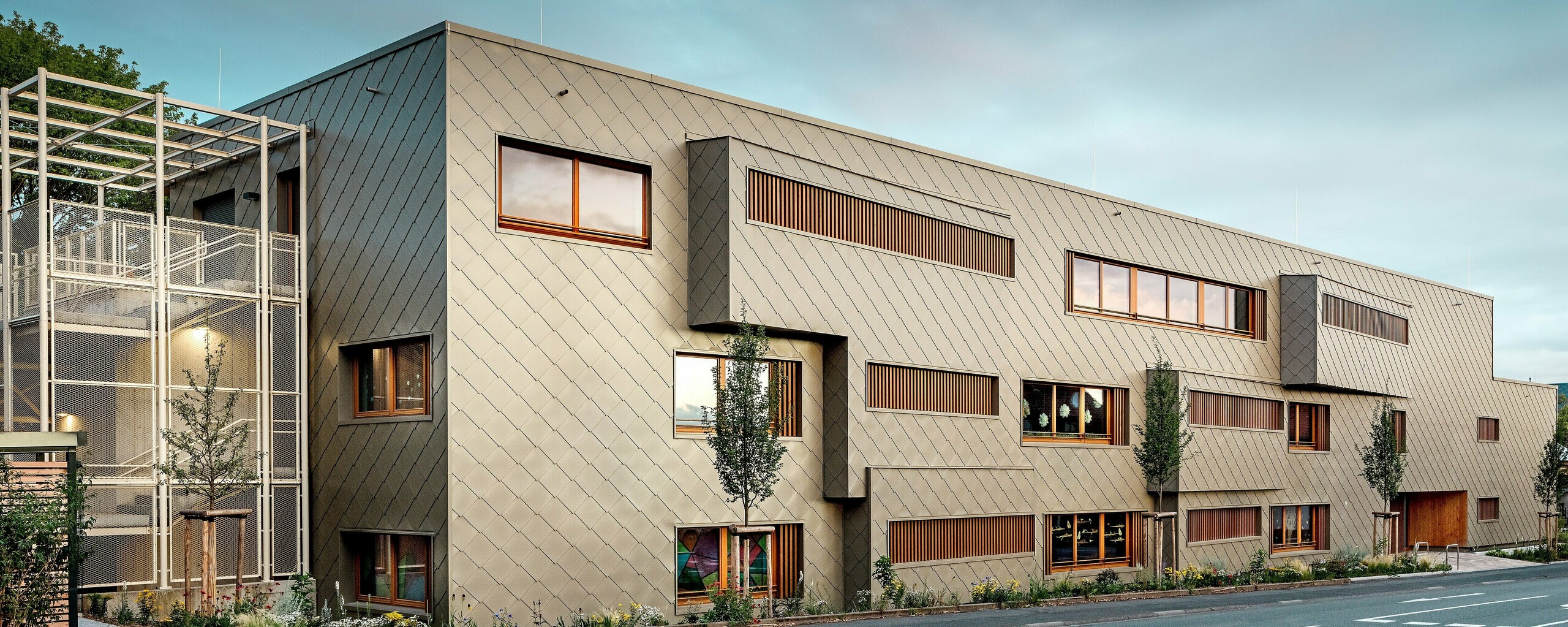 La photographie prise au niveau de la rue montre la nouvelle crèche d'Alzenau en dialogue dynamique avec le paysage urbain historique, notamment l'église remarquable en arrière-plan. La façade moderne, construite à partir de losanges de façade 44 x 44 couleur bronze, confère au bâtiment une élégance presque sculpturale. Des fenêtres judicieusement placées, des persiennes verticales en bois et des surfaces de façade décalées créent profondeur et rythme. À gauche, on aperçoit la cage d'escalier délicate avec sa structure métallique blanche, qui évoque une impression de légèreté moderne. La clarté du design, le choix de matériaux durables et la façade en aluminium de haute qualité font de ce bâtiment un projet phare d'intégration urbaine et de construction d'avenir grâce aux systèmes PREFA