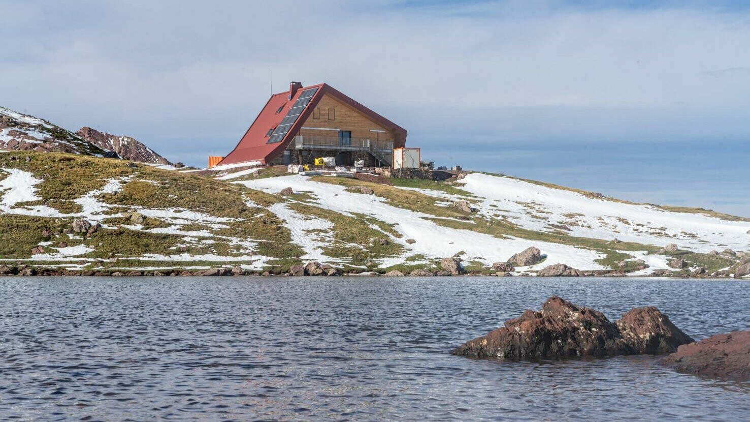 Vue d'ensemble du paysage : lac, montagne enneigé et le refuge d'Arlet rénové avec les losanges de toiture et de façade 44x44 PREFA dans la teinte rouge oxyde