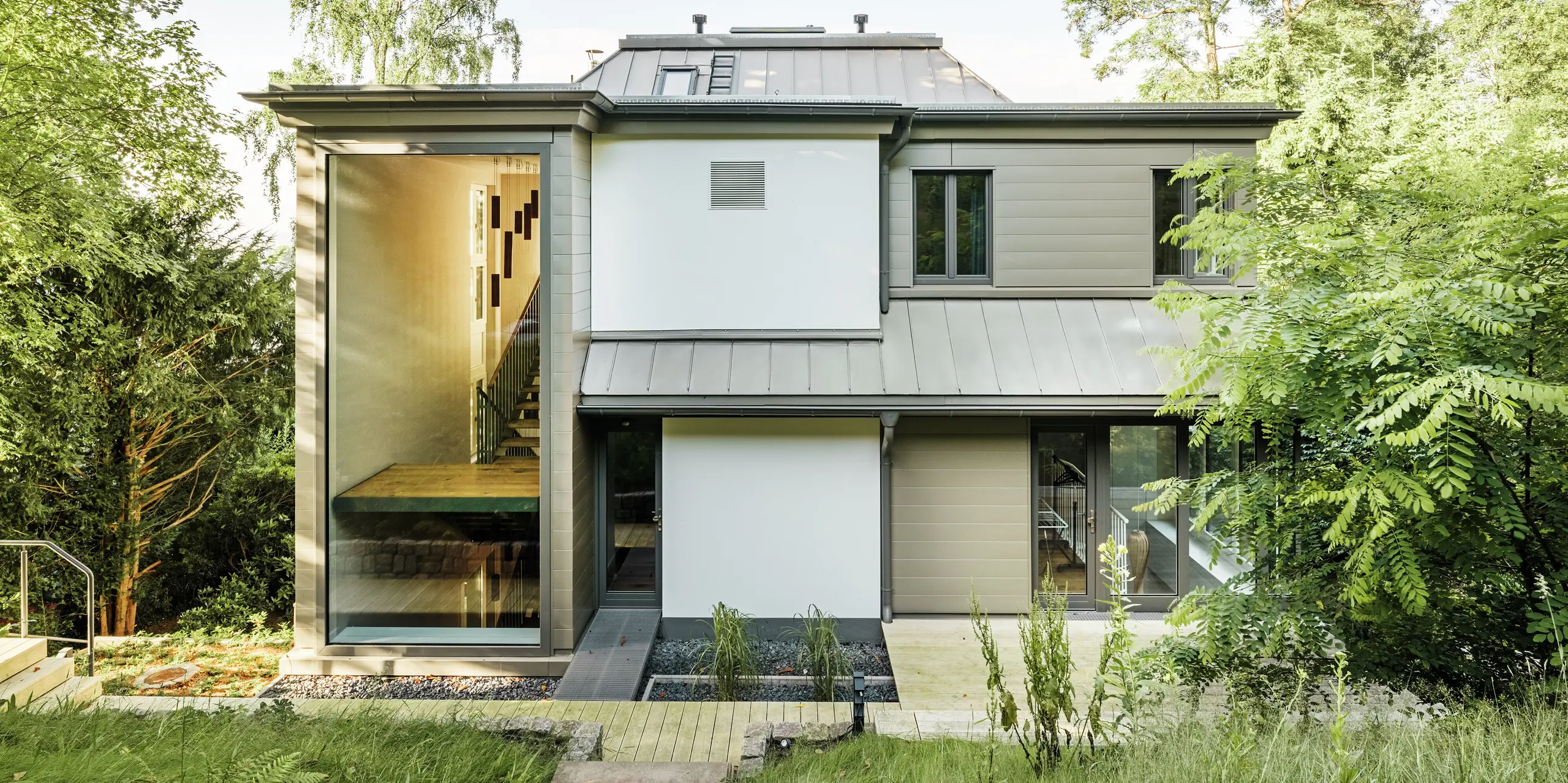 L'image présente la façade arrière de la Villa Blankenese à Hambourg, caractérisée par une architecture moderne et épurée. Le bardage PREFA couleur bronze est particulièrement remarquable et confère à la façade une élégance intemporelle. La toiture, revêtue de PREFALZ couleur bronze, s'intègre harmonieusement à l'ensemble. De larges surfaces vitrées et l'alliance de l'aluminium et du plâtre soulignent le caractère haut de gamme du bâtiment. Durables, les produits PREFA offrent non seulement un design raffiné, mais aussi une résistance maximale aux intempéries et ne nécessitent aucun entretien.
