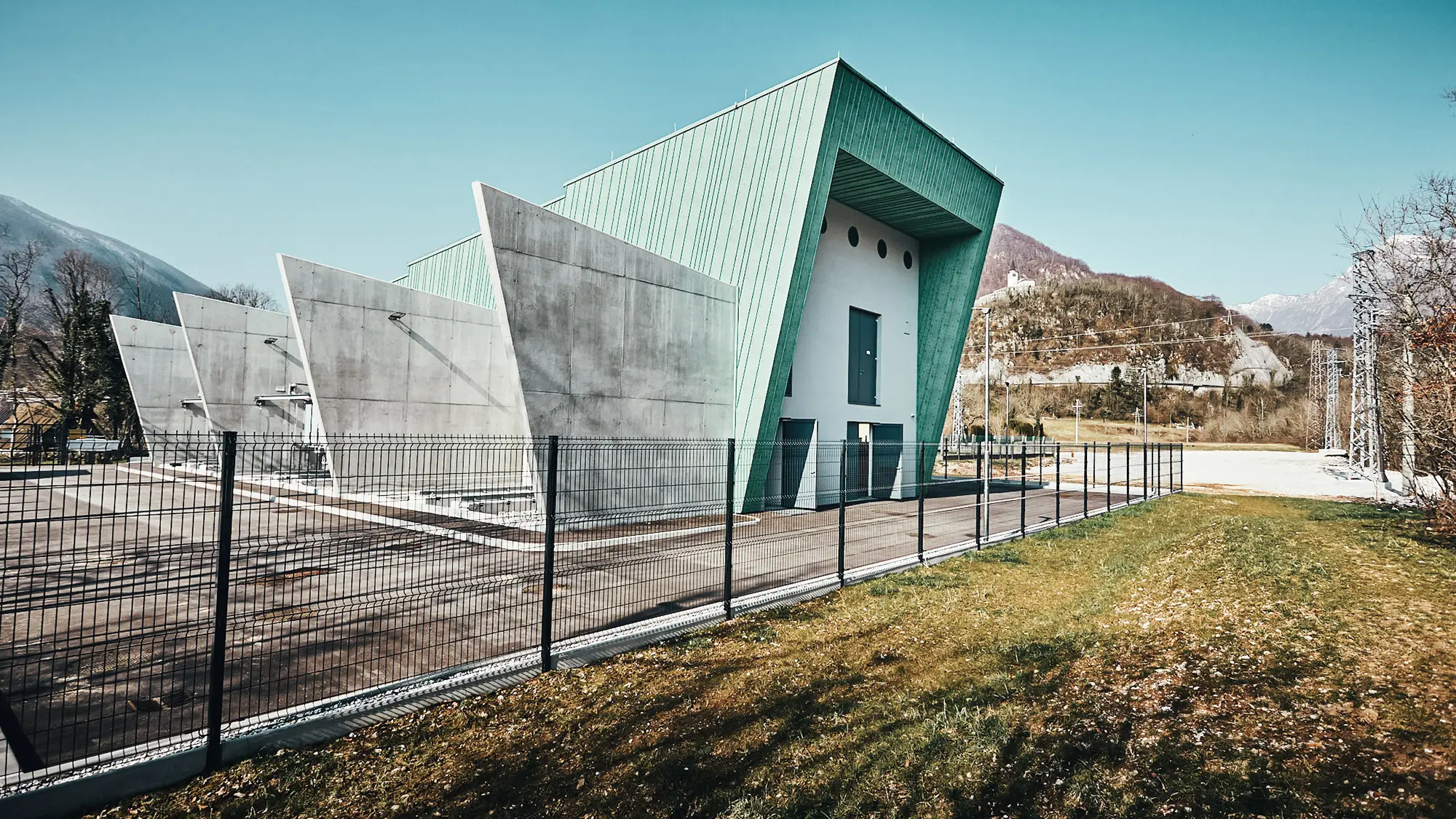 Seitenansicht des Umspannwerks samt Stirnseite und Betontrennwänden, eingezäunt und mit Bergen im Hintergrund.