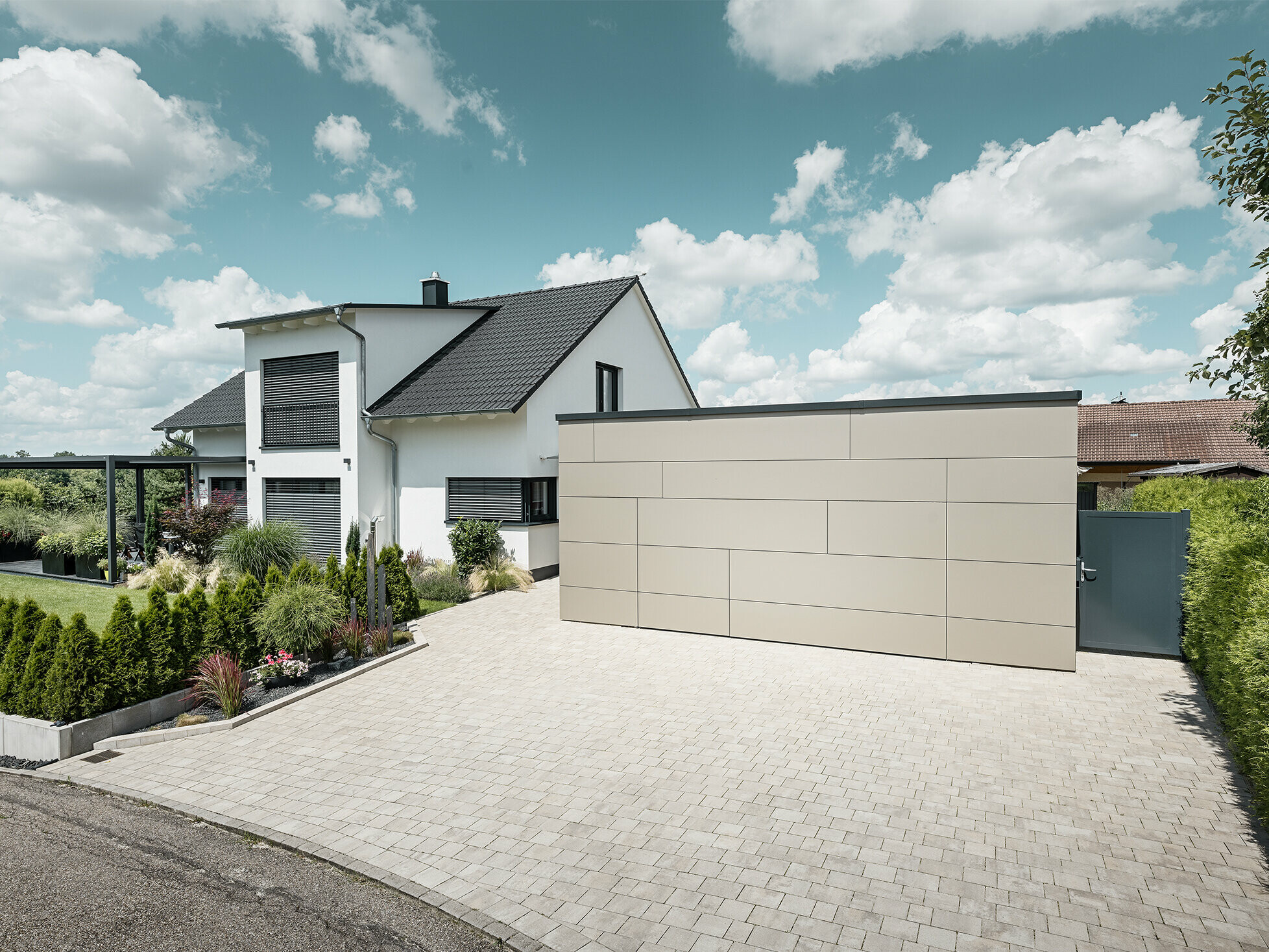 La façade du garage de cette maison individuelle est entièrement revêtue avec le panneau composite en aluminium couleur bronze. On ne distingue pas non plus la porte de garage des joints. Une allée spacieuse s'étend juste devant.