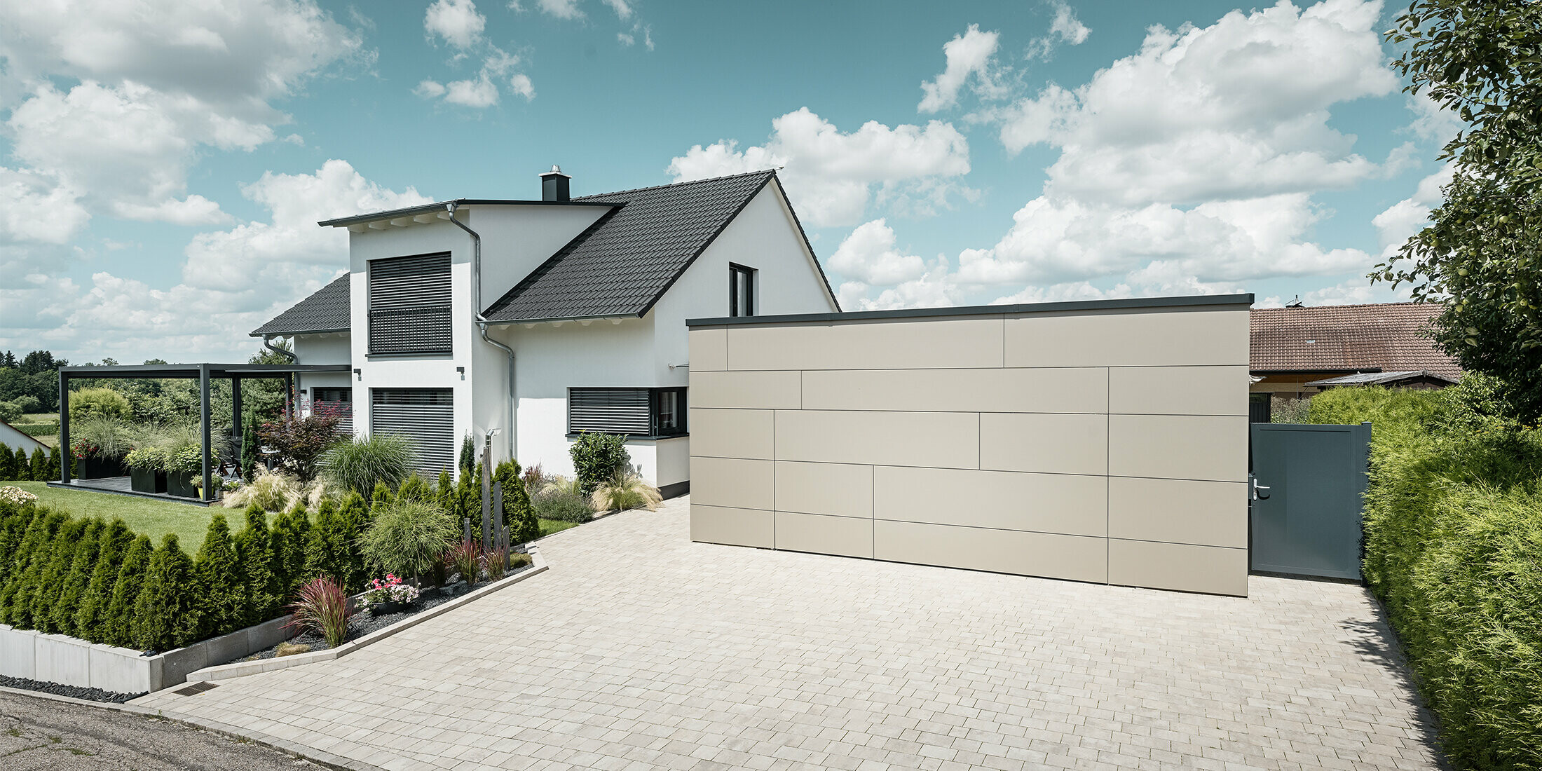 La façade du garage de cette maison individuelle est entièrement revêtue avec le panneau composite en aluminium couleur bronze. On ne distingue pas non plus la porte de garage des joints. Une allée spacieuse s'étend juste devant.
