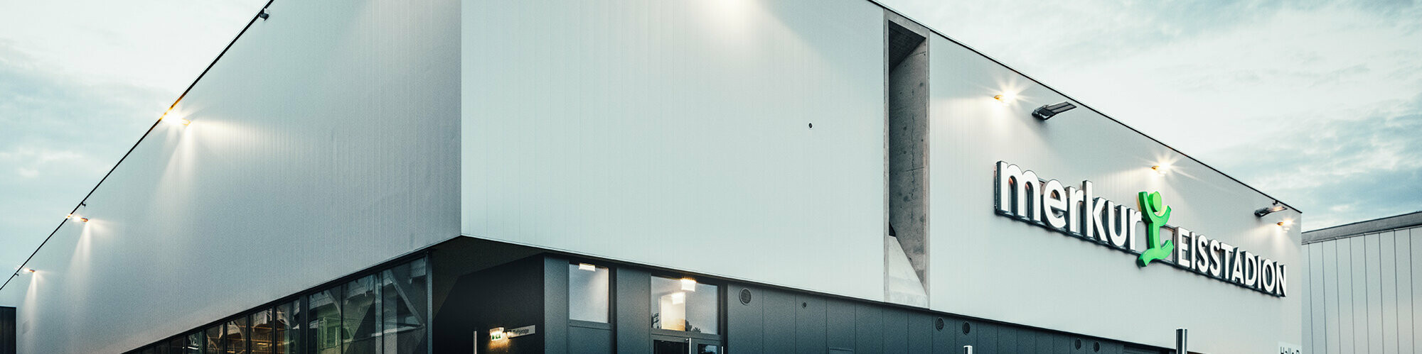 Prise de vue en angle de la patinoire. Sur le mur latéral droit, on peut voir une façade ventilée et l'inscription « merkur EISSTADION » sur la façade.