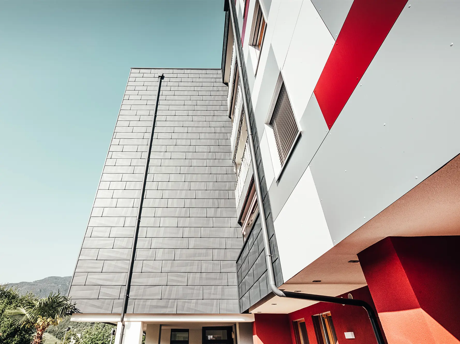 Prise de vue rapprochée et du dessous. En premier plan : la façade PREFA anthracite. A droite : le front de façade dans les teintes blanc, gris et rouge.