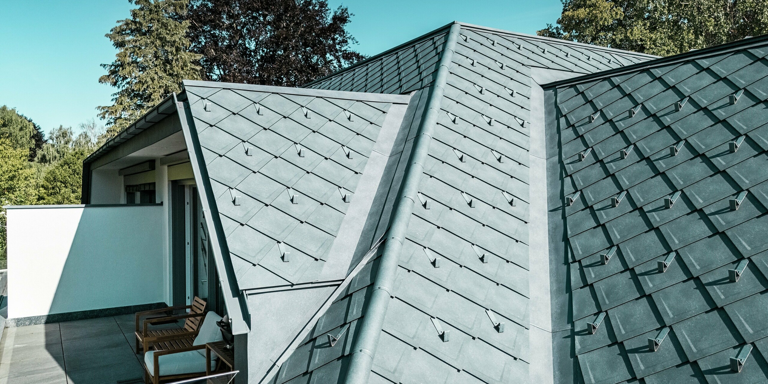 Vue détaillée d'une toiture PREFA en losanges 29 x 29 cm, coloris Gris Pierre P.10. L'image illustre la transition entre les pans de toiture et une élégante terrasse-balcon surplombant le toit. La pose précise des tuiles confère à la toiture une allure moderne et structurée. Les pare-neige qui l'ornent garantissent une sécurité accrue en hiver.