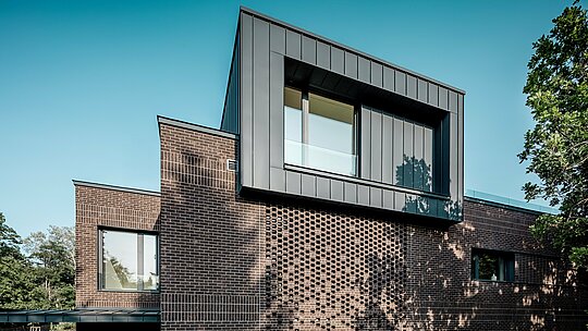 L'image révèle la façade remarquable d'une villa moderne multigénérationnelle à Budapest, où l'aluminium PREFALZ en P.10 anthracite affirme une identité architecturale forte. Le cube en porte-à-faux, avec ses joints debout angulaires précis, contraste fortement avec la façade en briques texturées. La lumière latérale du soleil crée un jeu dynamique d'ombres et de lignes, mettant en valeur la qualité d'exécution et la durabilité de la solution en aluminium PREFA. L'alliance du métal et de la brique souligne le caractère moderne et contrasté du bâtiment.
