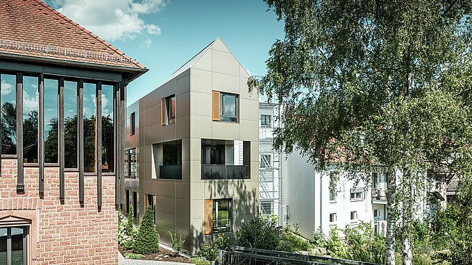 La photo montre la mairie moderne d'Alzenau, dont la façade est composée de panneaux composites en aluminium PREFA de couleur bronze. Le toit pointu et les fenêtres clairement structurées avec des stores en bois confèrent au bâtiment une esthétique élégante et contemporaine. Au premier plan, on aperçoit un espace vert avec un chemin pavé et des arbres qui complète harmonieusement le design urbain du bâtiment.