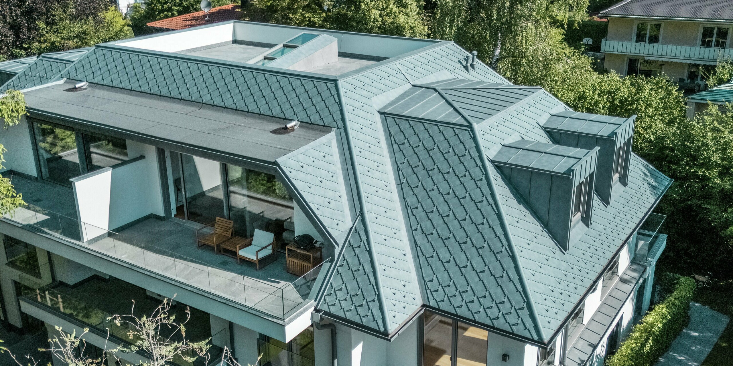 Vue aérienne d'un immeuble d'appartements moderne dont la toiture est recouverte de losanges PREFA 29 x 29, dans l'élégant coloris Gris Pierre P.10. La conception structurée et remarquable de la toiture est mise en valeur par de stylées lucarnes plates PREFALZ, parfaitement intégrées à l'architecture. Les généreuses terrasses et la végétation environnante renforcent l'impression d'élégance générale. L'utilisation de produits en aluminium de haute qualité garantit une solution durable et résistante aux intempéries, à la fois esthétique et fonctionnelle.