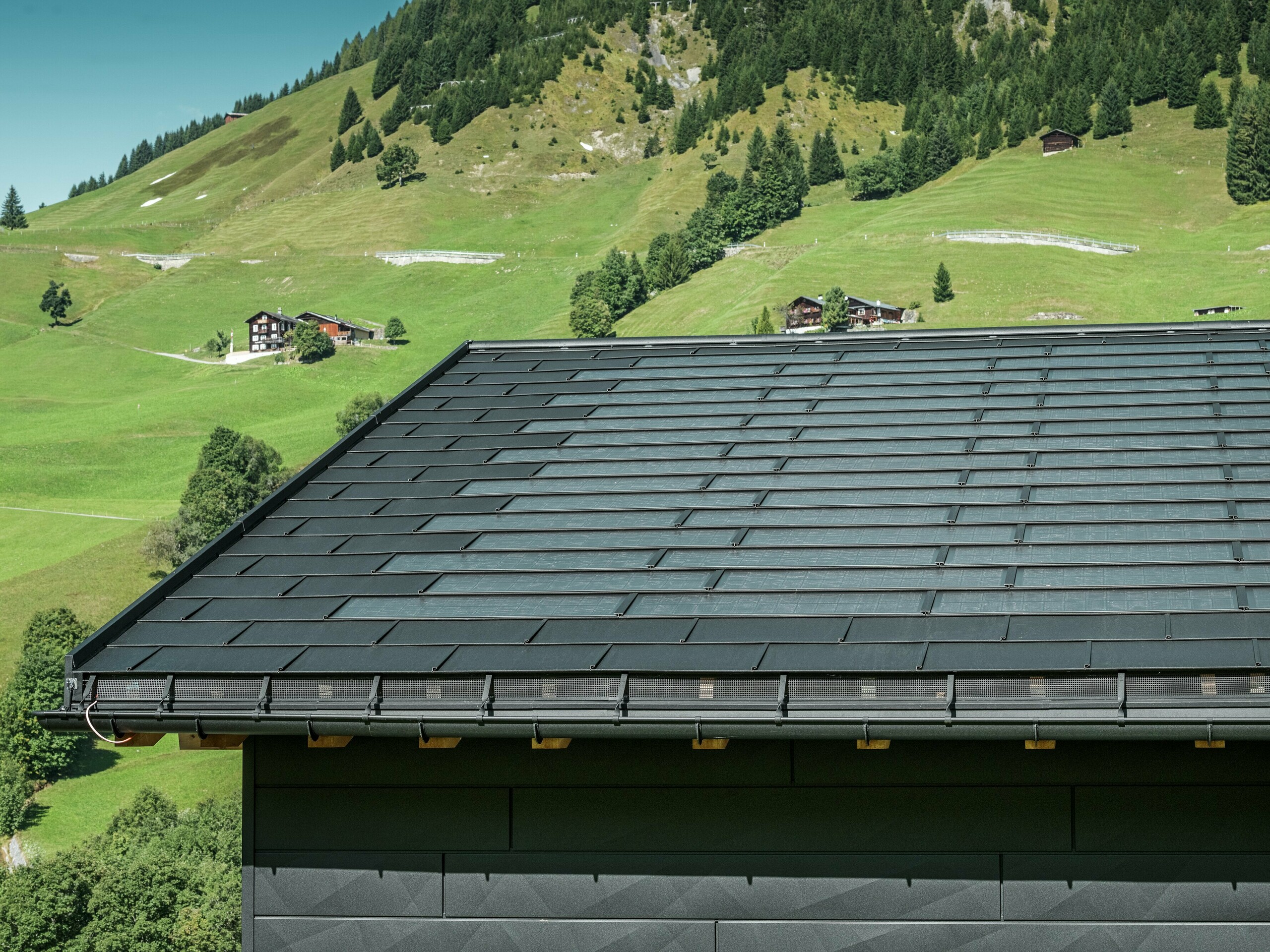 Gros plan sur l'avant-toit d'une maison familiale moderne dans les environs alpins d'Ascharina, en Suisse. Le toit a été recouvert de la tuile solaire innovante PREFA et du R.16 traditionnel, tous deux en P.10 noir. La gouttière suspendue PREFA et le tuyau de descente PREFA assurent un drainage fiable du toit. Les panneaux de façade distinctifs PREFA Siding.X sont visibles au bas de l'image. Les lignes claires des produits en aluminium soulignent le design moderne au milieu d'un paysage de montagne pittoresque.