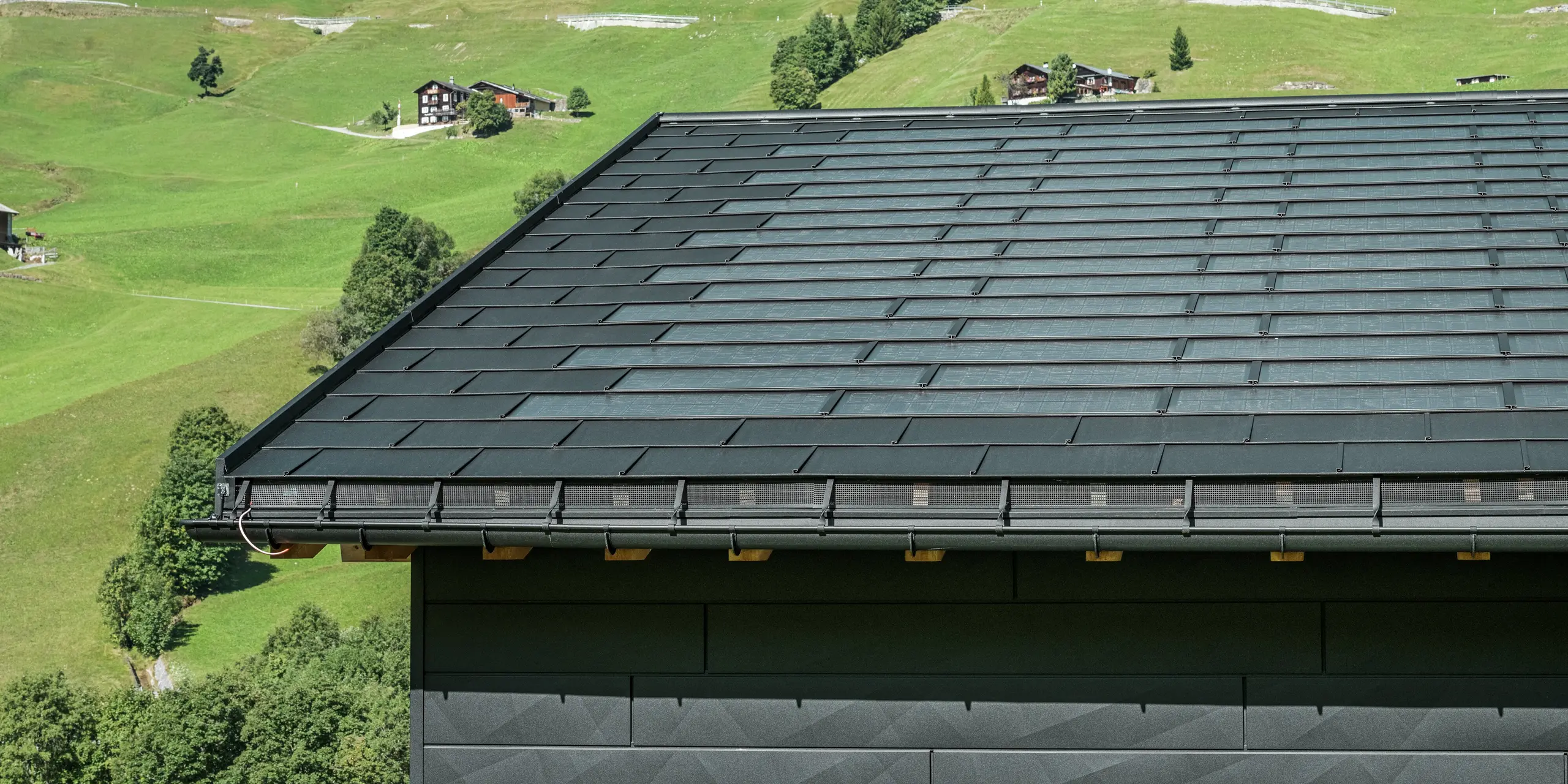 Gros plan sur l'avant-toit d'une maison familiale moderne dans les environs alpins d'Ascharina, en Suisse. Le toit a été recouvert de la tuile solaire innovante PREFA et du R.16 traditionnel, tous deux en P.10 noir. La gouttière suspendue PREFA et le tuyau de descente PREFA assurent un drainage fiable du toit. Les panneaux de façade distinctifs PREFA Siding.X sont visibles au bas de l'image. Les lignes claires des produits en aluminium soulignent le design moderne au milieu d'un paysage de montagne pittoresque.