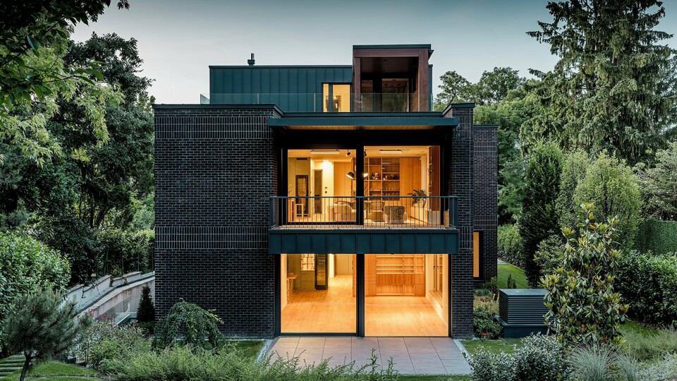 [Translate to Français:] Die Aufnahme zeigt die moderne Rückseite der Budapester Mehrgenerationen‑Villa in der Abenddämmerung. Großzügige Glasfronten öffnen den Blick auf warm erleuchtete Innenräume, während die Kombination aus dunkelroter Backsteinfassade und PREFALZ Aluminium in P.10 Anthrazit klare architektonische Linien betont. Die präzise ausgeführten Winkelstehfalze unterstreichen die hochwertige Verarbeitung und Langlebigkeit der PREFA Aluminiumprodukte. Umgeben von üppigem Grün fügt sich das Gebäude harmonisch in die Gartenlandschaft ein und vermittelt einen eleganten, modernen Charakter.