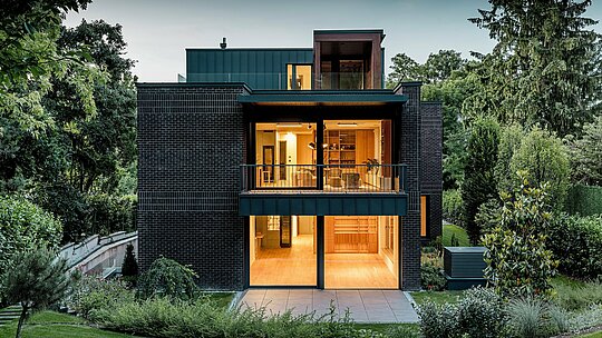 La photographie dévoile la façade arrière moderne de cette villa multigénérationnelle à Budapest, au crépuscule. De larges baies vitrées ouvrent sur des intérieurs baignés d'une lumière chaleureuse, tandis que l'alliance de la brique rouge foncé et de l'aluminium PREFALZ P.10 anthracite souligne la pureté des lignes architecturales. La précision de l'exécution des joints debout angulaires témoigne de la qualité de fabrication et de la durabilité des produits en aluminium PREFA. Entouré d'une végétation luxuriante, le bâtiment s'intègre harmonieusement au jardin et affirme un caractère élégant et contemporain.