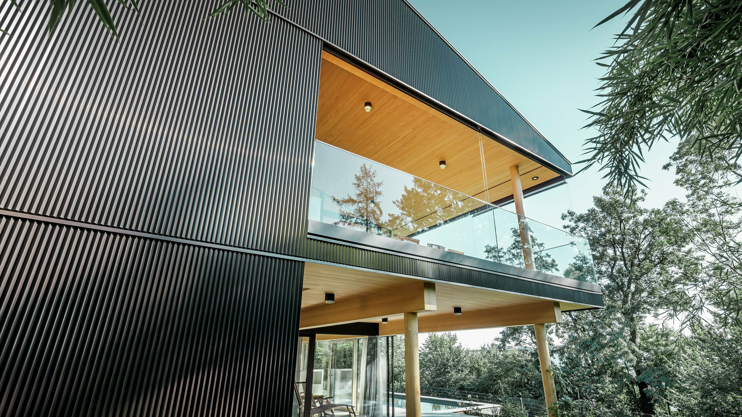 Vue latérale d'une maison individuelle, la façade est revêtue d'un profil triangle PREFA foncé. On aperçoit la terrasse et un grand balcon. Le plafond et la sous-face sont habillés de panneaux de bois.