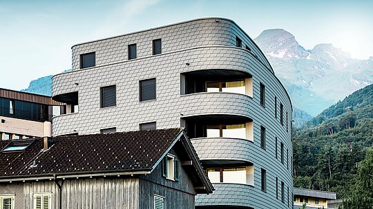 À Buchs, un immeuble résidentiel et commercial triangulaire et moderne se distingue par son architecture arrondie et sa façade en bardeaux, réalisée en bardage PREFA gris clair P.10. L'enveloppe en aluminium, parfaitement circulaire, lui confère une allure reptilienne dynamique et attire élégamment le regard sur le fond de montagnes alpines. Grâce à la légèreté, la résistance aux intempéries et la durabilité de l'aluminium PREFA, même les rayons de courbure les plus serrés sont exécutés avec précision, offrant ainsi une enveloppe homogène et facile d'entretien, au charme résolument moderne.