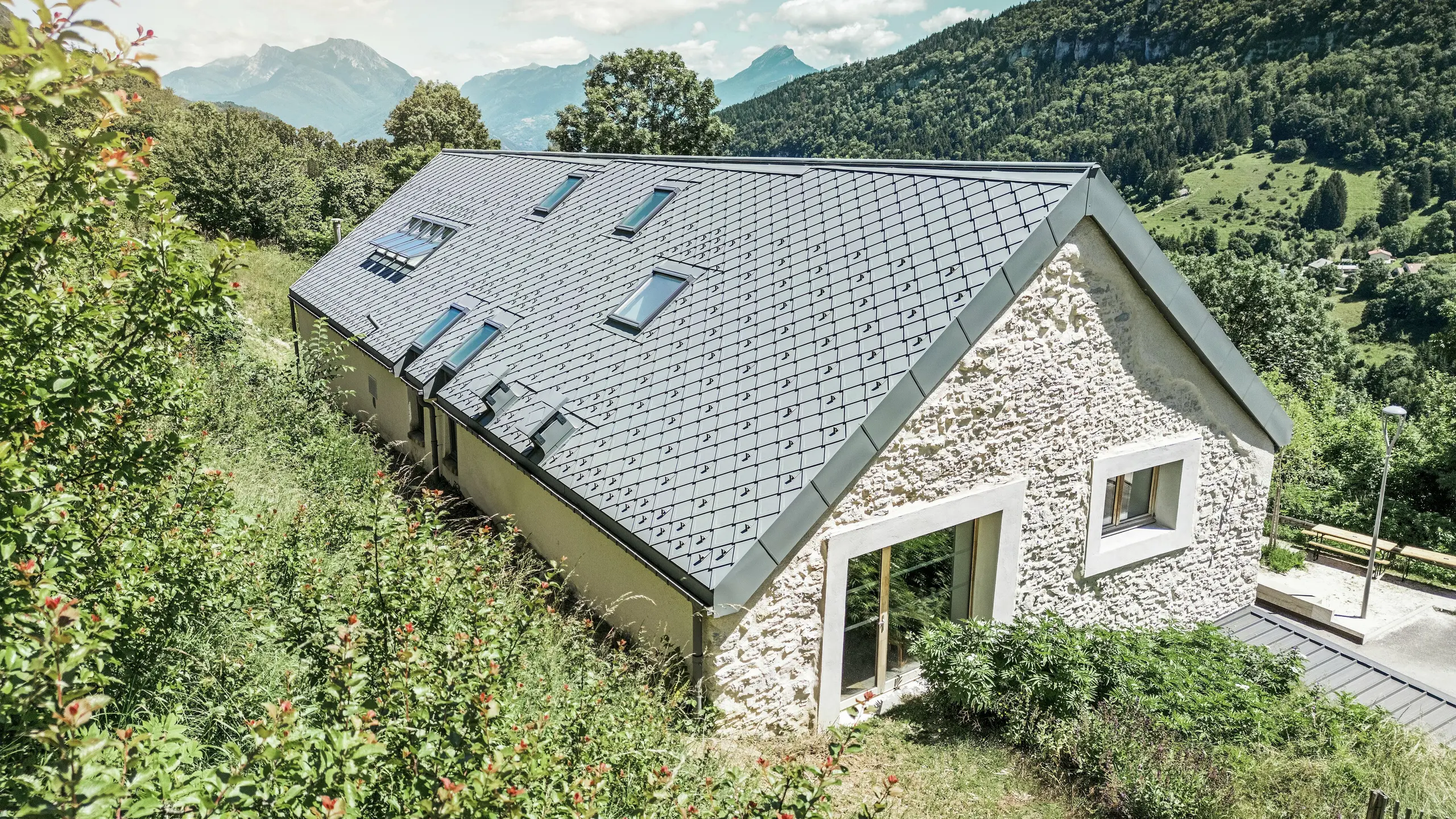 Vue du ciel de la ferme rénovée "La Grange à Gaby" à Engins, France. Le bâtiment est recouvert de losanges de toiture PREFA 29 × 29 durables, qui combinent une protection moderne avec une esthétique traditionnelle. Le paysage de montagne idyllique souligne l'intégration harmonieuse dans la nature.