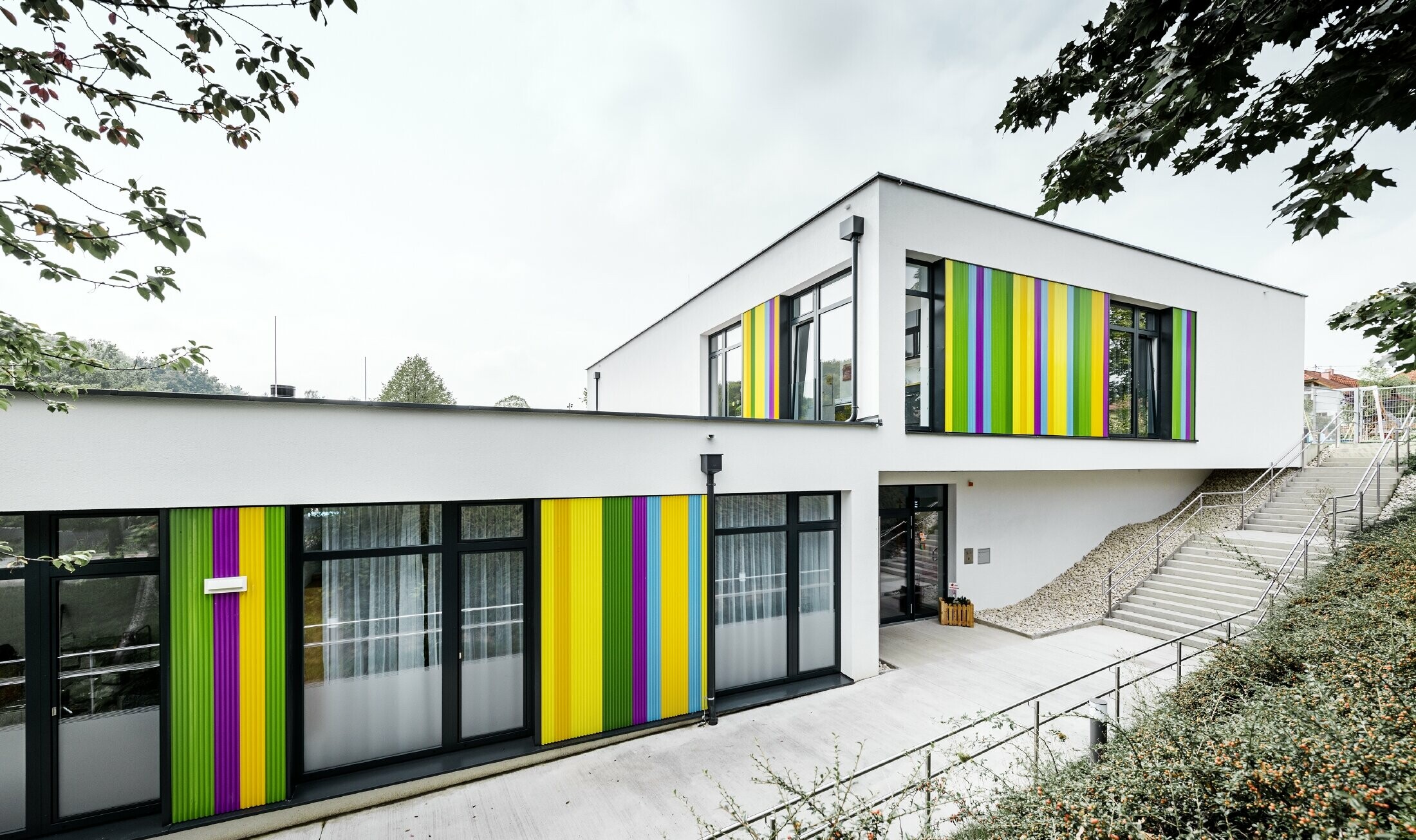 Éléments de façade multicolores de PREFA pour le jardin d’enfants de Hargelsberg. Le bâtiment présente un toit plat et des grandes fenêtres au niveau du sol.