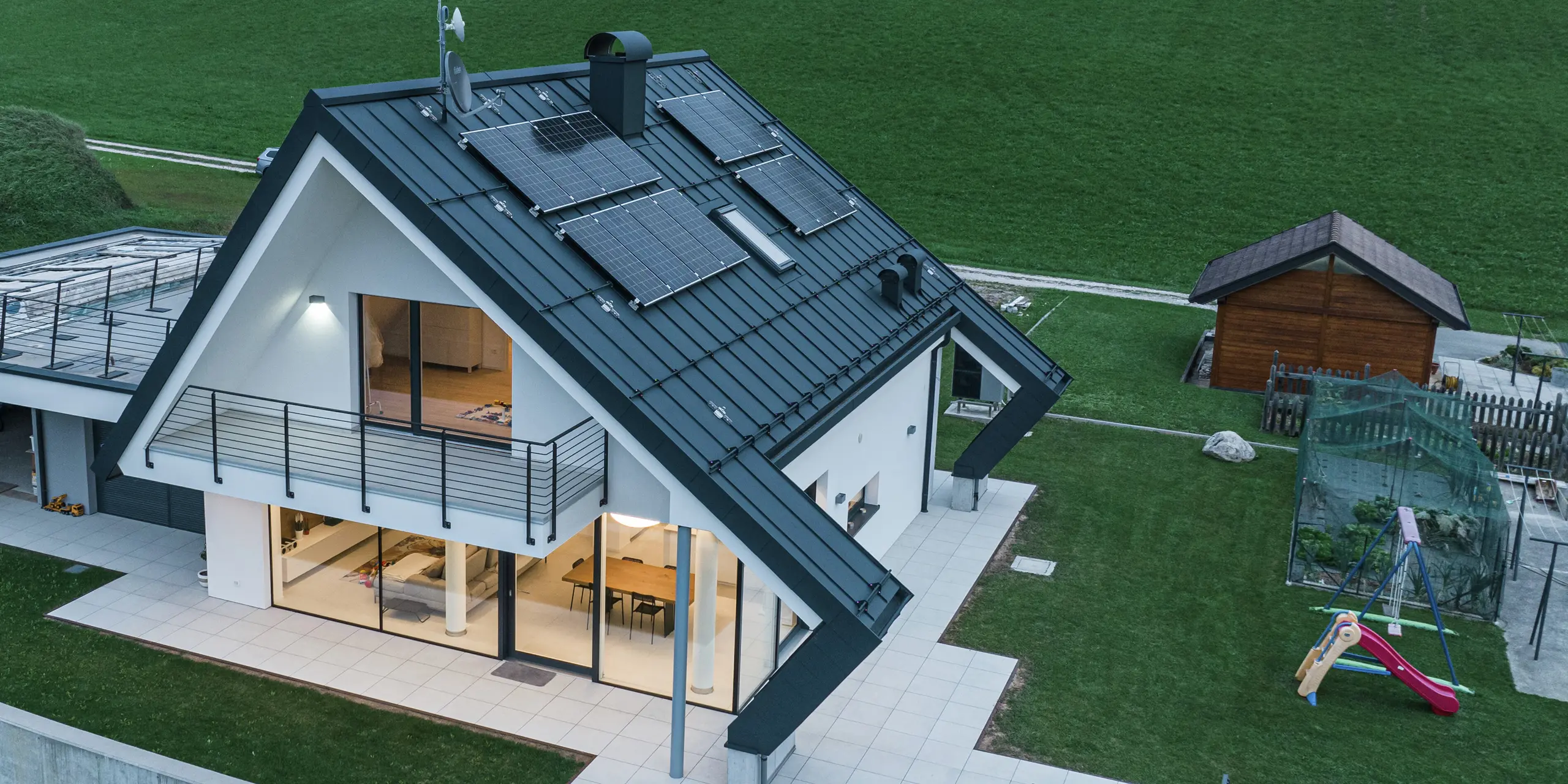 Maison individuelle moderne à Borgo Valbelluna vue à vol d'oiseau au crépuscule. Dans la maison, des lumières illuminent les pièces à vivre. Le toit remarquable avec ses surplombs inhabituels a été recouvert d'une toiture à joint debout PREFALZ robuste en P.10 anthracite. Le toit en aluminium durable et les modules photovoltaïques allient durabilité et design de haute qualité. Le surplomb du toit remarquable protège la façade et la terrasse des intempéries. De grandes surfaces vitrées créent une atmosphère de vie lumineuse, tandis que le balcon enveloppant et la terrasse spacieuse maximisent le confort de vie. L'aluminium PREFA est synonyme de stabilité, d'élégance et de fonctionnement sans entretien.