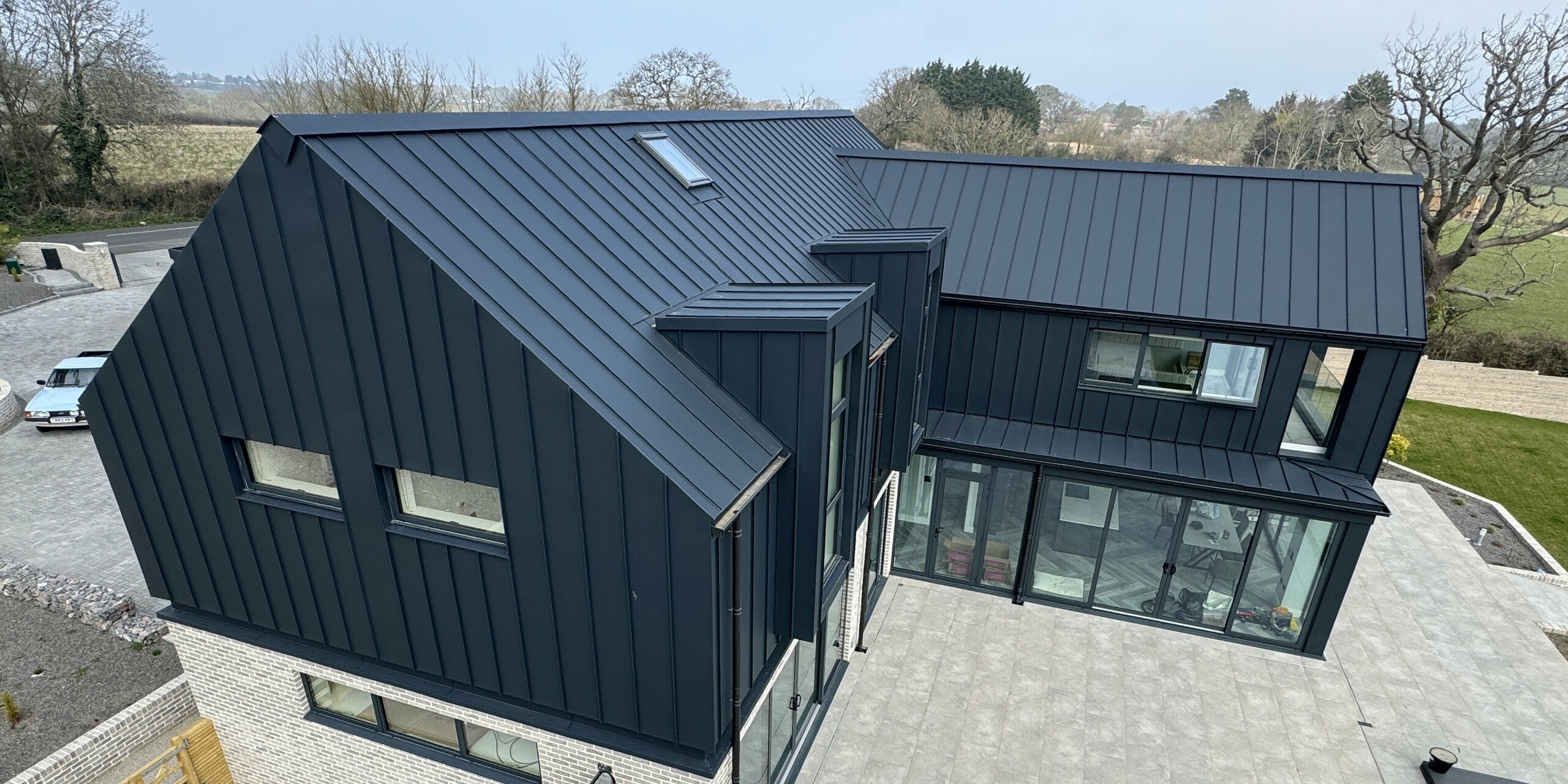 Vue détaillée d'une maison individuelle moderne avec une toiture et une façade PREFALZ en P.10 gris sombre. La forme originale de la toiture, avec ses joints debout précis, et les larges surfaces vitrées définissent le design. Au rez-de-chaussée, un revêtement en pierre claire contraste avec la façade en aluminium à joints debout foncés. La spacieuse terrasse en dalles grises encadre le bâtiment et offre une vue dégagée sur le jardin. PREFALZ séduit par sa durabilité, sa résistance à la corrosion et son absence d'entretien – un atout majeur pour l'exposition du site sur l'île de Wight.