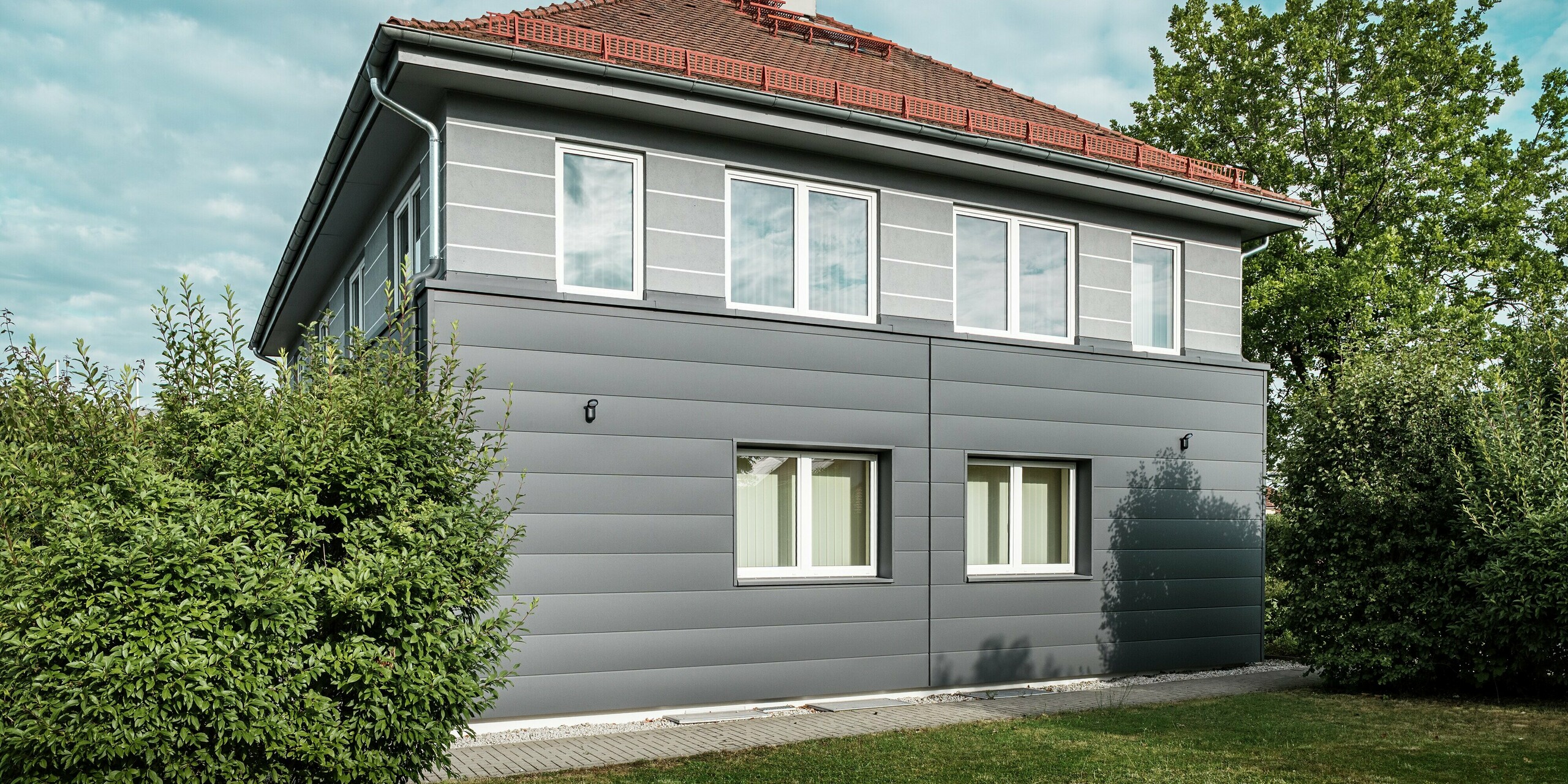 L'image présente la façade récemment rénovée de la guilde des ramoneurs à Bindlach, revêtue de Siding PREFA moderne dans l'élégant coloris Gris sombre P.10. Les panneaux d'aluminium à la structure apparente confèrent au bâtiment une allure contemporaine et stylée, tout en le protégeant efficacement des intempéries. La surface durable et nécessitant peu d'entretien est particulièrement remarquable, garantissant ainsi la préservation de l'état impeccable de la façade. L'alliance de la fonctionnalité et du design moderne fait du bardage PREFA un choix idéal pour les projets de rénovation exigeants.