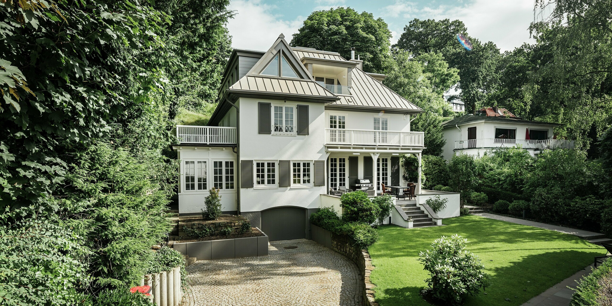 L'image présente la Villa Blankenese à Hambourg, dotée d'un vaste jardin et d'une élégante allée. La toiture PREFALZ couleur bronze, au design saisissant, définit l'architecture et crée des lignes épurées et modernes. La technique de la toiture à joints debout offre non seulement une esthétique raffinée, mais aussi une résistance maximale aux intempéries. Les lucarnes et les éléments de façade sont revêtus de Siding PREFA couleur bronze, soulignant ainsi l'harmonie entre la toiture et la façade. L'alliance de l'aluminium durable et d'un design classique fait de cette villa une référence en matière d'architecture sophistiquée.