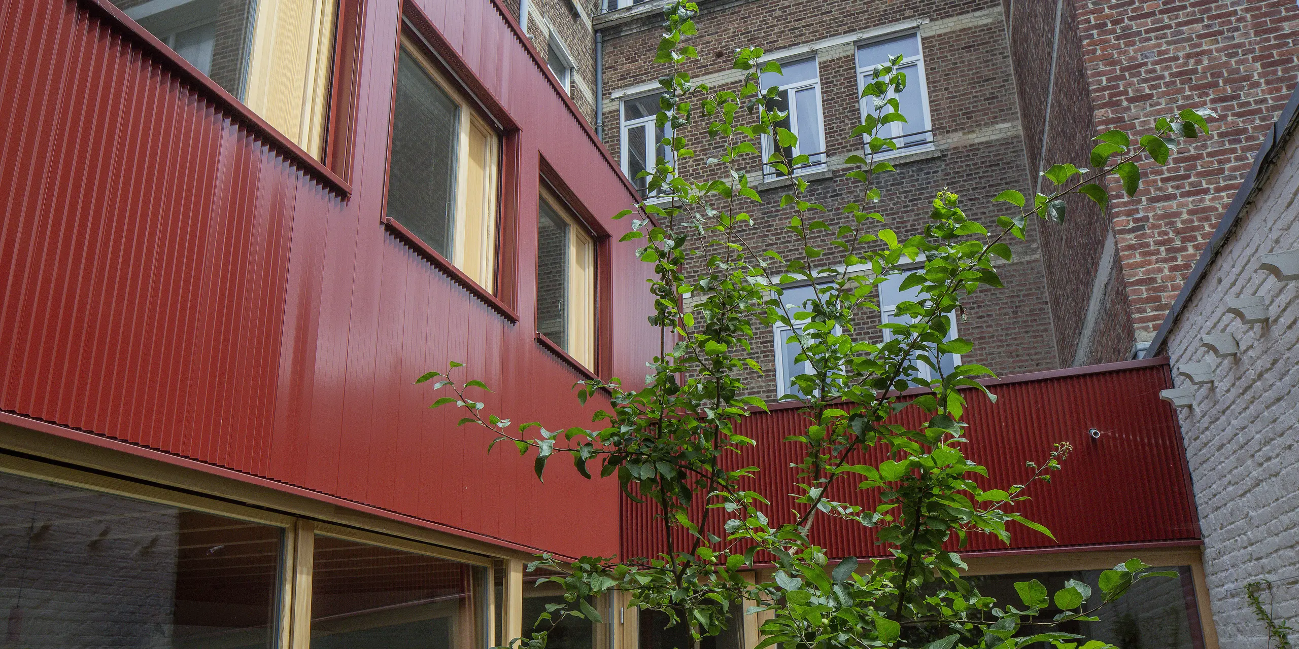 Du vert sur du rouge : au premier plan, un arbre se dresse, encadré par les lignes épurées du profil triangle PREFA rouge oxyde. La façade structure et donne de la profondeur à la cour urbaine, créant une atmosphère moderne et agréable. Le revêtement en aluminium, sans entretien, résiste aux intempéries et au temps, et constitue un atout architectural durable.
