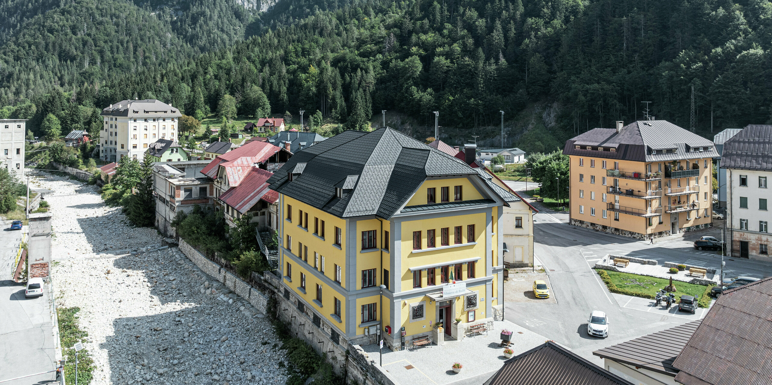 Niché dans un paysage de montagne impressionnant se trouve le Museo Cave del Predil en Italie. Le musée avec sa façade jaune frappante est équipé de tuiles PREFA de haute qualité de couleur P.10 anthracite. Le toit en aluminium durable garantit une protection optimale contre les conditions météorologiques alpines telles que la neige et les tempêtes, associée à un design esthétique. En arrière-plan, le panorama alpin s'élève majestueusement avec des forêts denses et des pics rocheux.