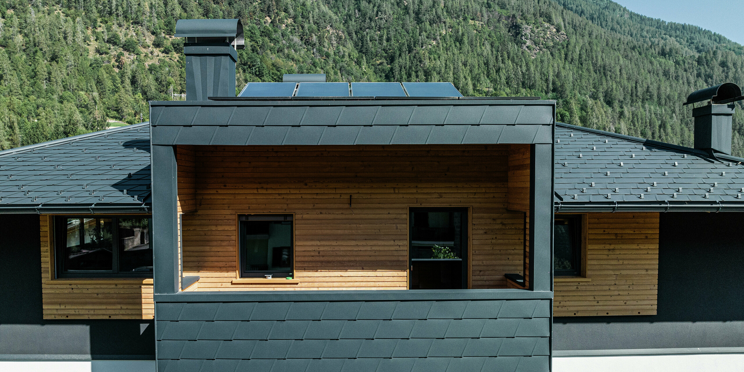 Gros plan sur les DS.19 PREFA en P.10 anthracite sur le toit d'un immeuble résidentiel à Commezzadura, Italie. Le revêtement en aluminium assure un design moderne et durable sur le toit et la façade des balcons couverts. La façade en bois naturel sous le revêtement en tôle foncée crée une impression générale harmonieuse.