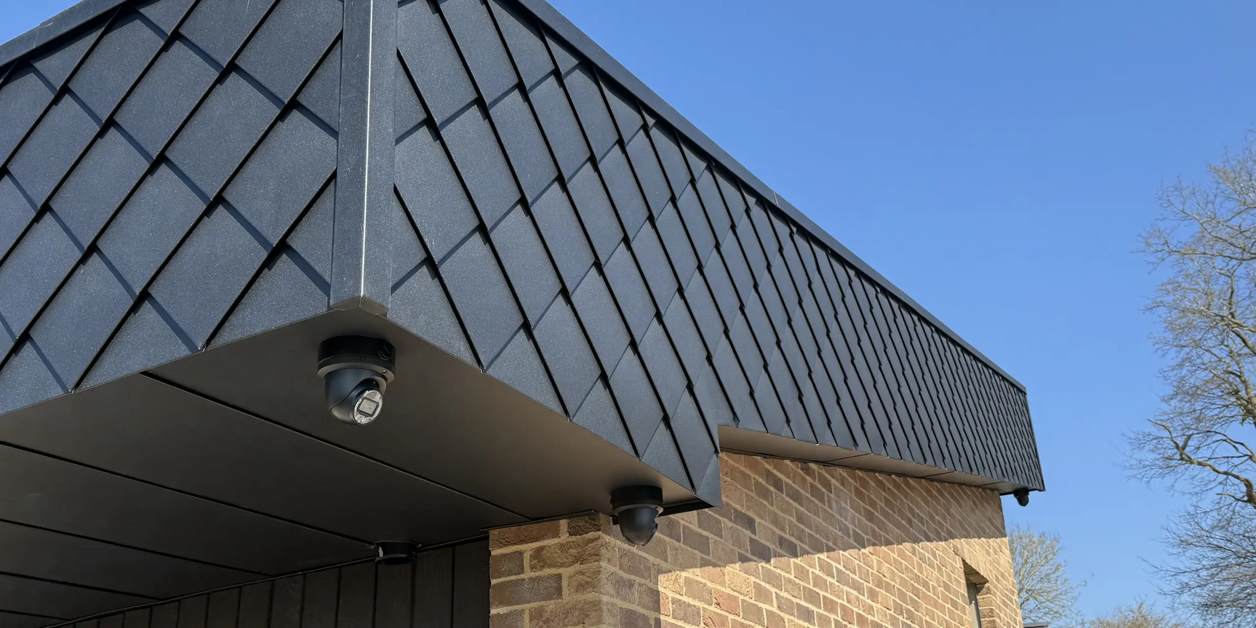 Vue détaillée d'une façade moderne du projet résidentiel « Doone Brae Farm », caractérisée par une transition saisissante entre la brique clinker chaleureuse et les élégants losanges de façade PREFA 20 × 20 en P.10 anthracite. Ces panneaux en aluminium créent un effet visuel dynamique et soulignent la pureté des lignes du bâtiment. Le contraste entre la brique clinker naturelle et l'aluminium de haute qualité confère au design un caractère intemporel et une grande efficacité énergétique.