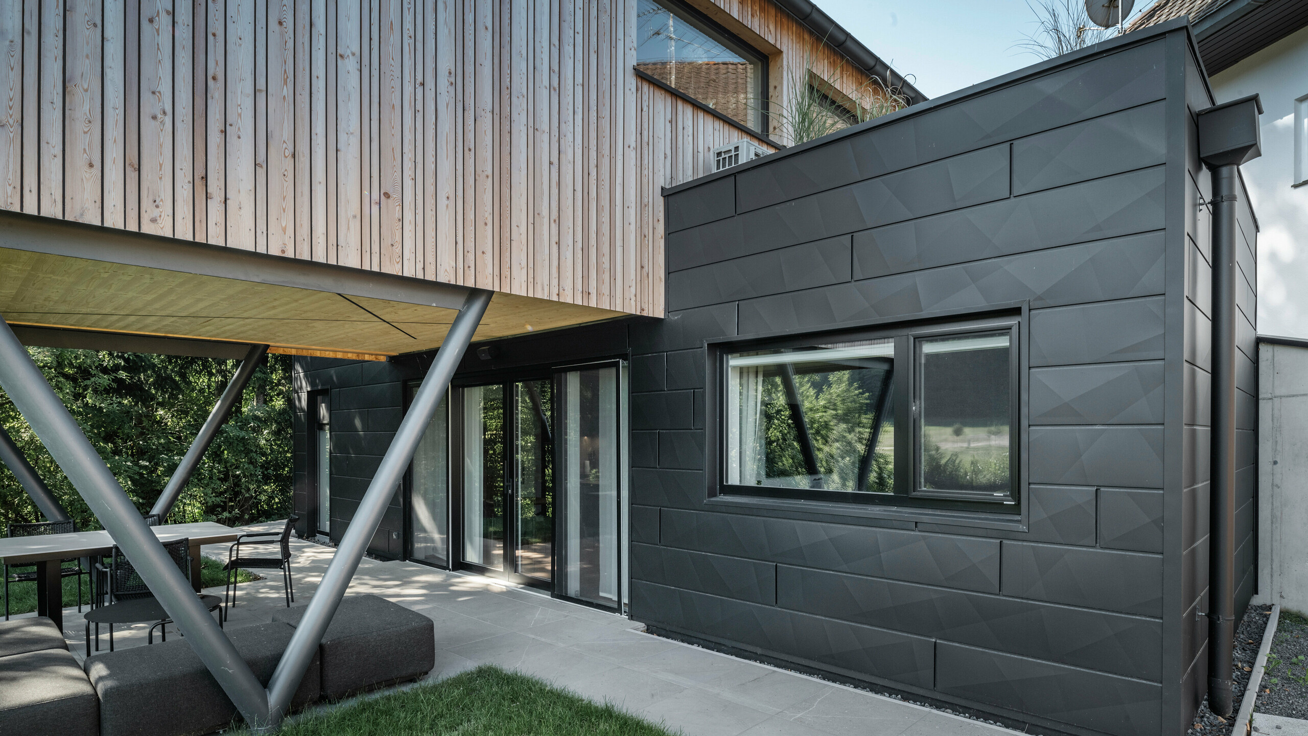 Joyau architectural : « Woodstock Chalet 5 » à St. Martin am Innkreis, en Haute-Autriche. Ce lieu moderne dédié aux mariages est mis en valeur par l'utilisation de panneaux de façade PREFA Siding.X de couleur anthracite. Les éléments en aluminium contrastent fortement avec les panneaux en bois clair et les structures métalliques de soutien. Les cadres de fenêtres sombres s'intègrent harmonieusement dans la conception de la façade, conférant au bâtiment un aspect élégant. Cette image montre l'équilibre esthétique entre la texture naturelle du bois et le charme industriel de la façade en aluminium, qui caractérise la conception des bâtiments contemporains.