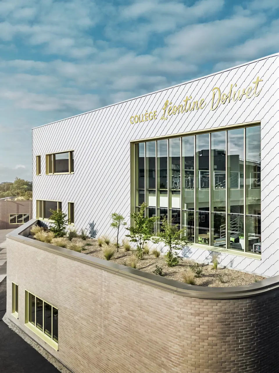 La photo révèle un angle remarquable du collège Léontine Dolivet à Cesson-Sévigné. La façade est composée de losanges PREFA argent métallisé, agencés en diagonale et jouant avec la lumière. La forme incurvée de l'étage supérieur est particulièrement frappante et confère au bâtiment une allure douce et moderne. De larges baies vitrées offrent des vues sur l'intérieur, tandis que les encadrements de fenêtres jaunes apportent des touches de dynamisme. Au premier plan, un espace planté de gravier et d'arbustes s'intègre harmonieusement à l'architecture. Le ciel légèrement couvert accentue encore la brillance des surfaces métalliques.