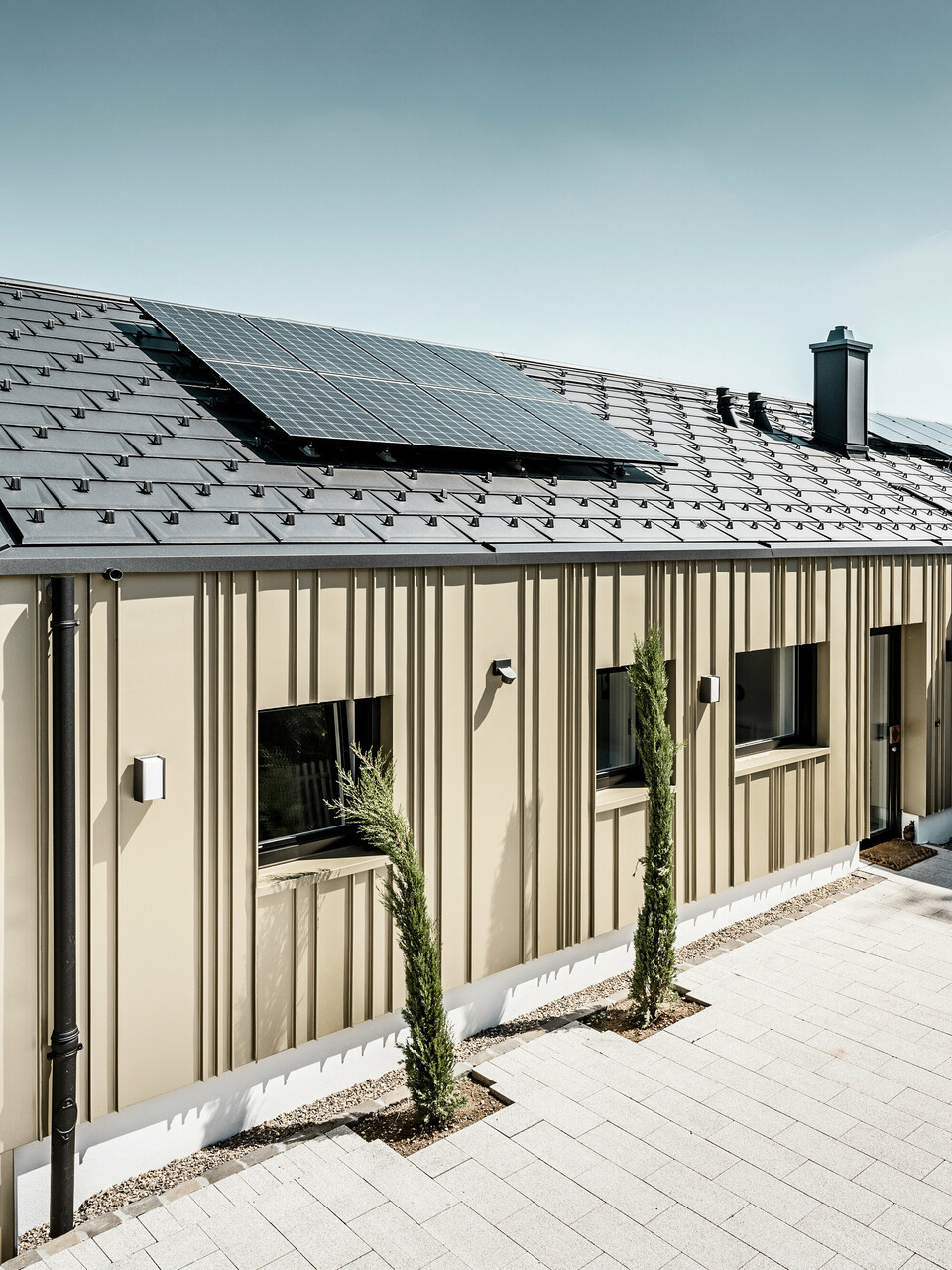 Architektonisch ansprechendes Einfamilienhaus, harmonisch in die Hügellandschaft eingebettet, mit einem hochwertigen PREFA Dachsystem in Form der Dachplatte R.16 in elegantem P.10 Anthrazit. Die Fassade zeichnet sich durch eine geschmackvolle Winkelfalzkonstruktion aus Falzonal in Lichtbronze aus, die das Gebäude mit einem warmen, metallischen Glanz umhüllt. Das Haus verfügt zudem über eine umweltfreundliche Komponente in Form von Solarmodulen, die diskret auf dem Dach integriert sind. Die PV-Anlage sowie die Gebäudehülle aus langlebigem Aluminium betonen das Engagement für nachhaltiges Bauen. Eine minimalistische Garage fügt sich nahtlos an das Hauptgebäude an, während die gepflasterte Einfahrt und sorgfältig platzierte Grünpflanzen das Bild eines modernen, ökologisch bedachten Zuhauses vervollständigen.