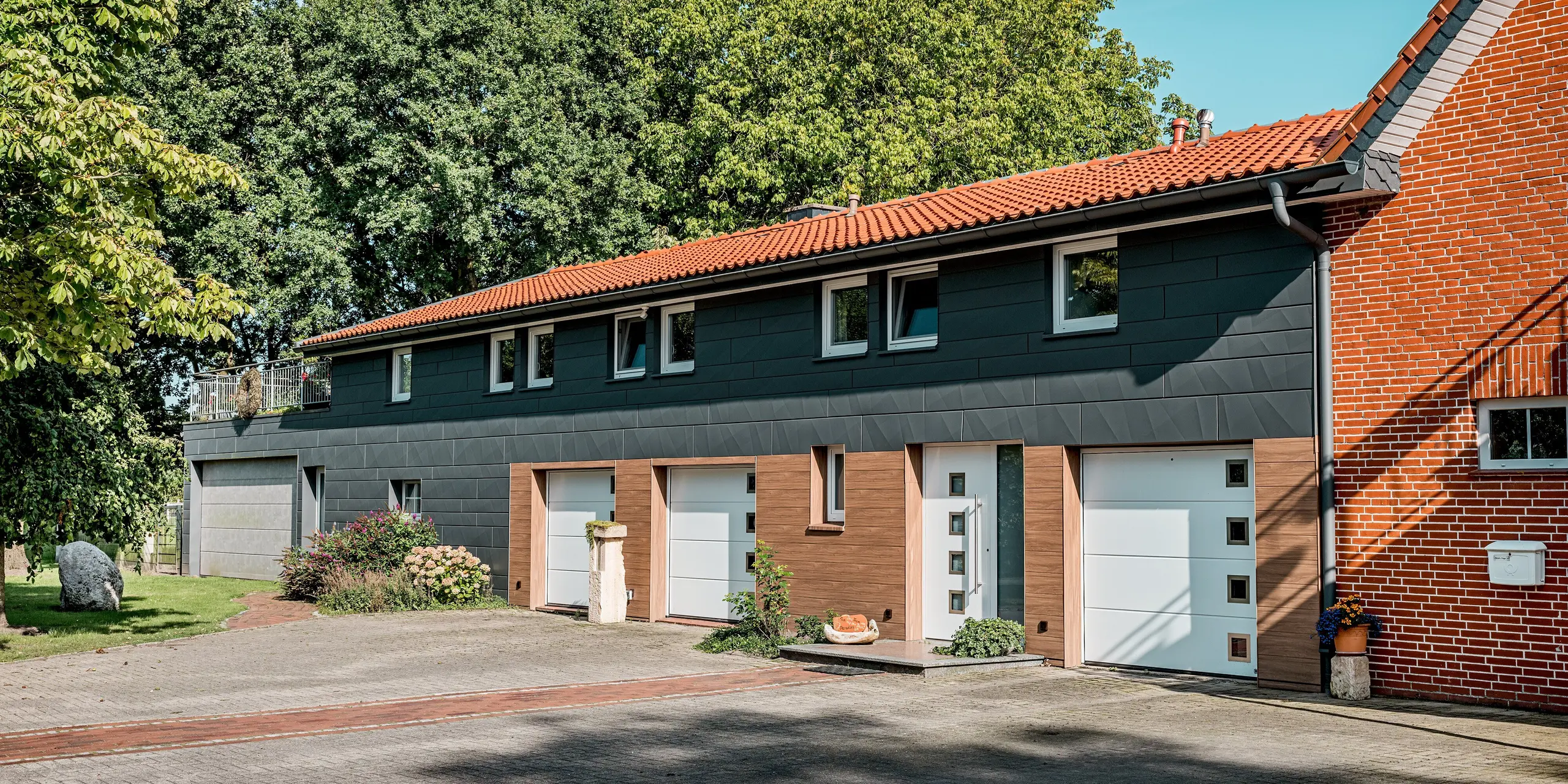 Cette maison individuelle, rénovée avec goût, arbore une façade en aluminium PREFA au design horizontal remarquable. Parmi ses atouts, on note l'alliance du bardage PREFA, d'un chaleureux aspect noyer foncé, au rez-de-chaussée et à l'entrée, et du bardage Siding.X, couleur P.10 gris sombre à l'étage. La netteté des contours des panneaux foncés crée de captivants jeux d'ombre et de lumière, conférant à la façade une élégance contemporaine. Les portes de garage blanches et les fenêtres aux contours bien définis apportent un contraste harmonieux. Durables, résistants aux intempéries et faciles d'entretien, les panneaux en aluminium PREFA sont idéaux pour les rénovations durables, alliant design moderne et grande fonctionnalité.