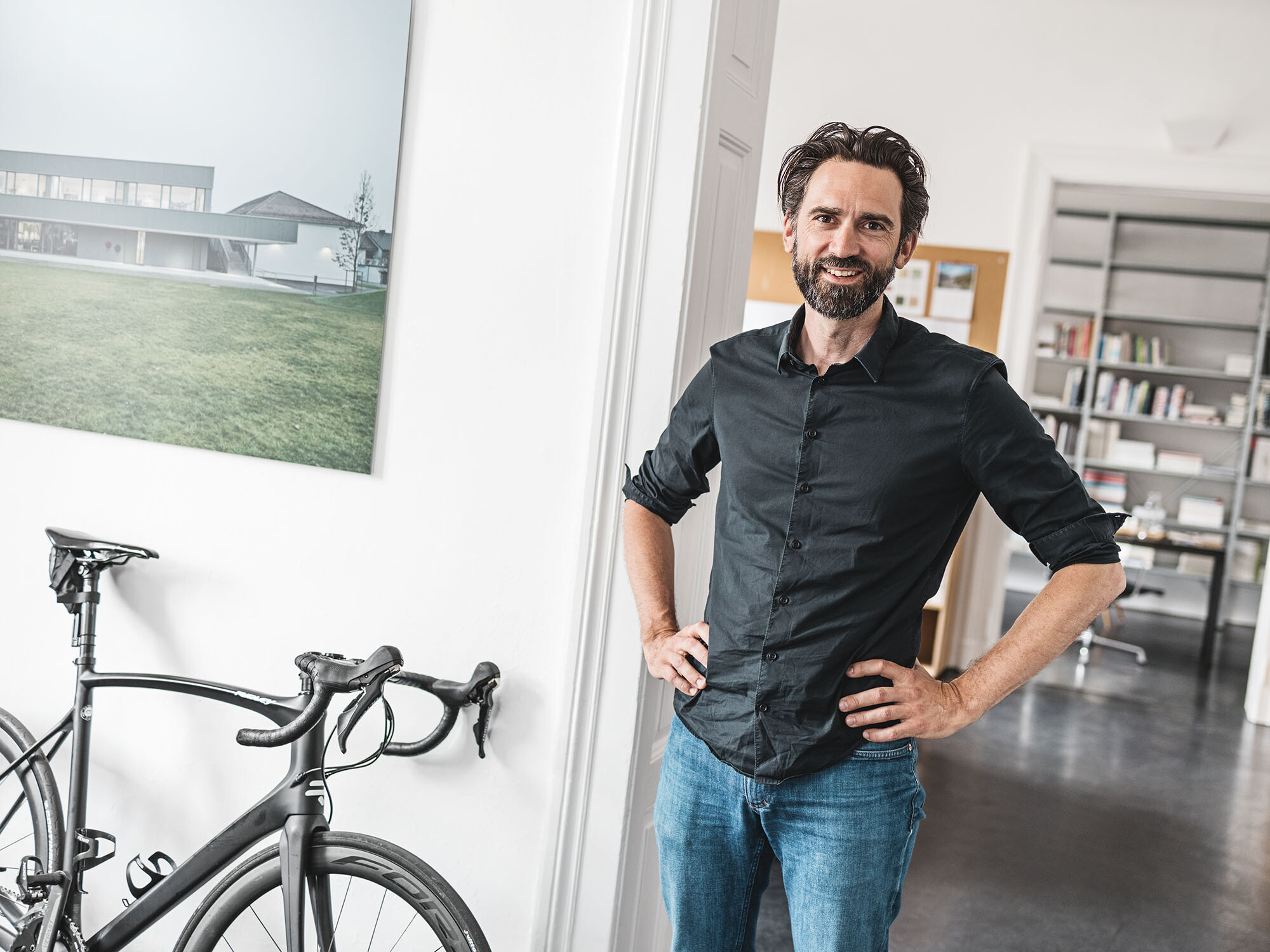 Portrait de l'architecte en chef Thomas Heil ; derrière lui s'étend un espace de bureau, à côté de lui, un vélo est appuyé contre le mur.