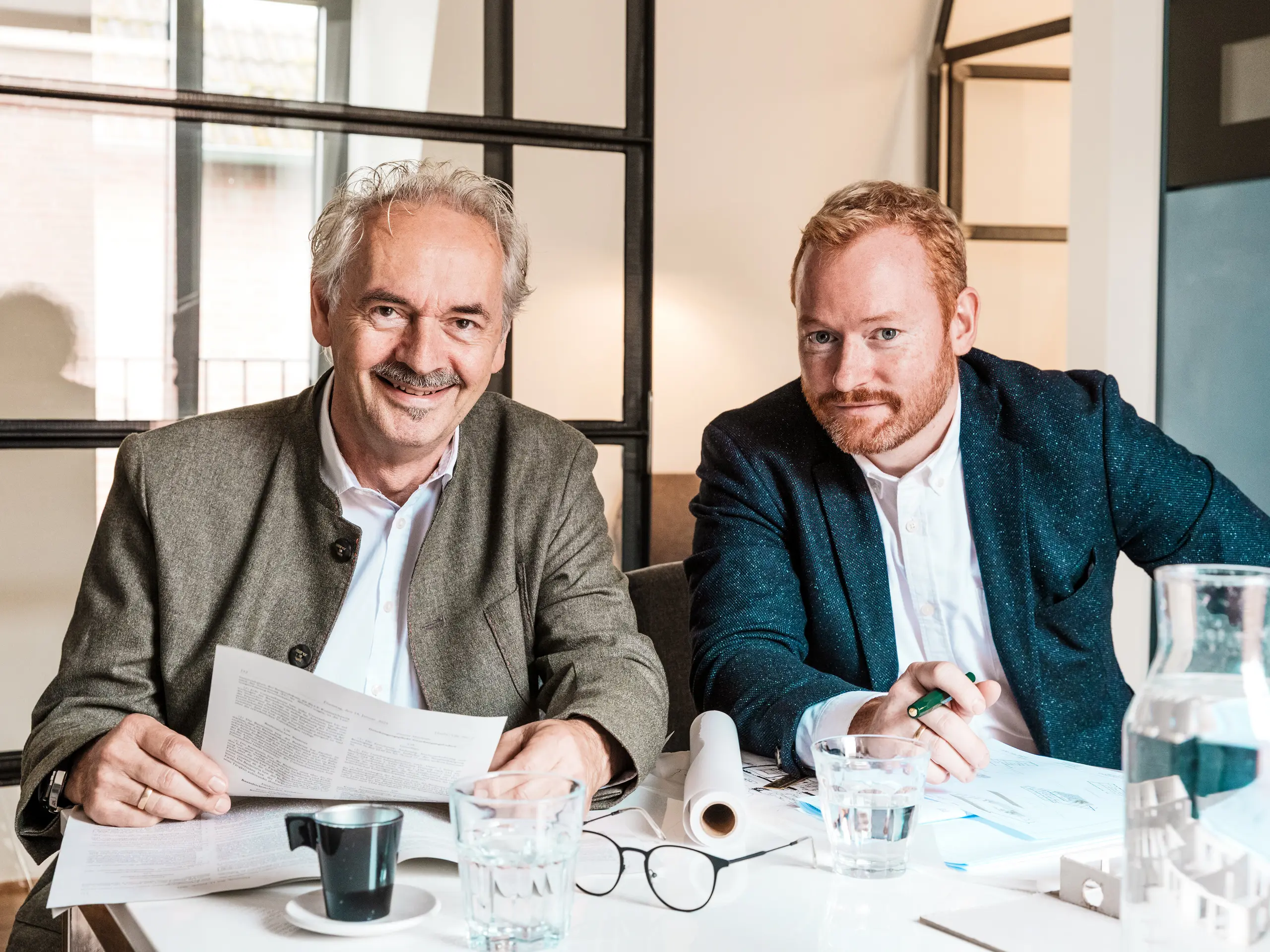 Deux architectes sont assis à une table, en train de dessiner des plans. Des boissons et une maquette sont posées devant eux. Le commissariat 43 de Hambourg, conçu par les deux architectes, a été agrandi de façon remarquable grâce à l'utilisation de profils extrudés PREFA. La scène met en valeur les deux hommes du cabinet d'architectes Scheffczyk Architekten. L'image illustre leur travail, à travers les plans, les maquettes et les documents, témoignant du long processus qui a mené à l'achèvement du bâtiment.