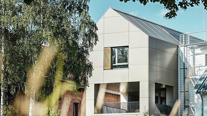 La photo montre la mairie moderne d'Alzenau, dont la façade est composée de panneaux composites en aluminium PREFA dans un élégant ton bronze. Le toit pointu et les grandes fenêtres soulignent le caractère contemporain du bâtiment. Au premier plan, des arbres et des espaces verts encadrent le bâtiment, tandis que l'éclairage doux et le ciel clair soulignent l'espace urbain moderne.