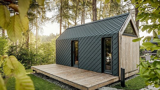 Nichée dans un écrin de verdure paisible au cœur du Secret Koi Garden, sur les rives du lac Natterer, une minuscule maison minimaliste arbore un toit et une façade revêtus de losanges en aluminium PREFA P.10 anthracite. Leur structure précise, évoquant des écailles de poisson, reflète délicatement la lumière du soleil. La spacieuse terrasse en bois ouvre l'espace de vie sur la nature environnante, tandis que l'aluminium PREFA, résistant aux intempéries et durable, protège la structure et crée un contraste saisissant avec le bois et la végétation.