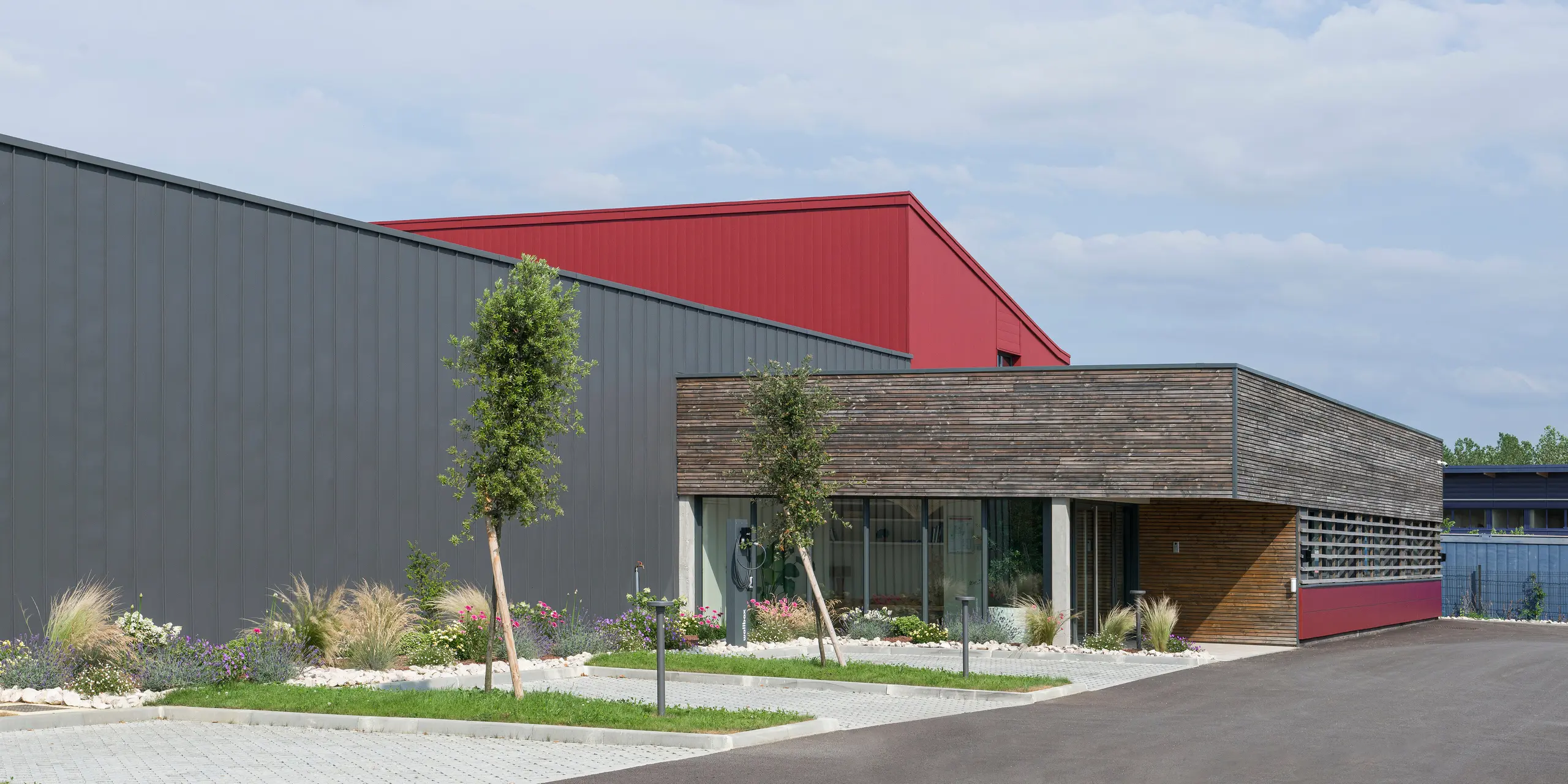 L’image montre un bâtiment moderne avec une combinaison frappante d’éléments de design industriel et naturel. La façade combine des éléments en aluminium gris foncé et rouge oxyde qui apportent dynamisme et structure. Les revêtements PREFA rouge oxyde apportent une touche colorée. La partie basse du bâtiment, dotée d'un mur revêtu de bois et de grandes fenêtres, crée une atmosphère chaleureuse et invitante, évoquant un espace à usage de bureau. Le jardin bien entretenu devant le bâtiment véhicule une image respectueuse de l'environnement et durable. La combinaison de ces éléments reflète le désir de combiner des méthodes de construction traditionnelles avec des matériaux et des conceptions modernes et durables.